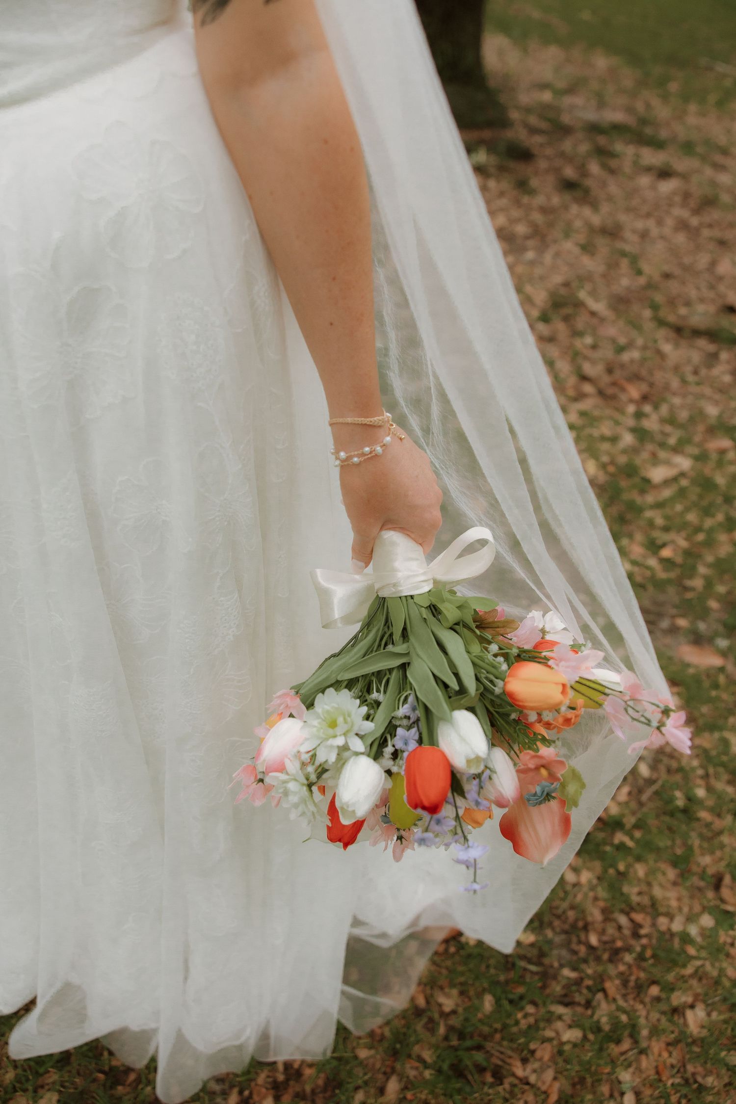 A bride in white wedding dress holds a colorful spring bouquet with tulips and pink flowers against her flowing veil.