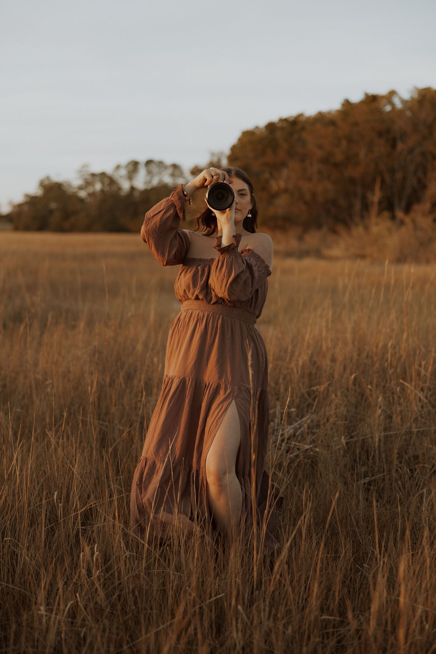 A photographer in a flowing brown dress captures photos in a golden field at sunset.