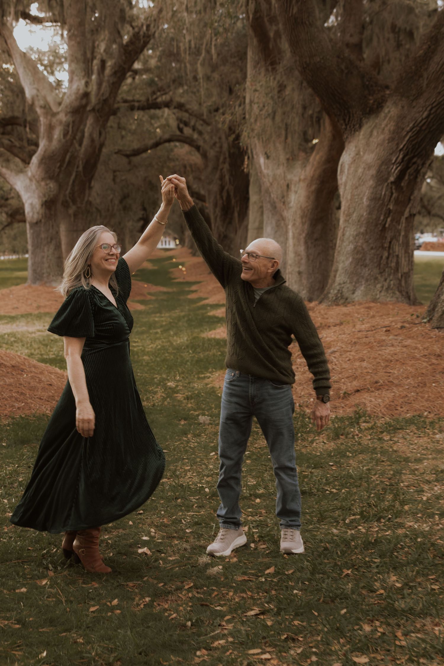 Dancing couple in casual attire joyfully holding hands under a canopy of large trees.
