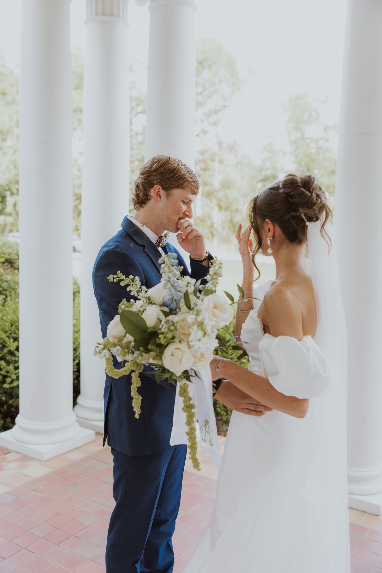 Bride and groom share a tender moment during their first look in an elegant white-columned outdoor setting.
