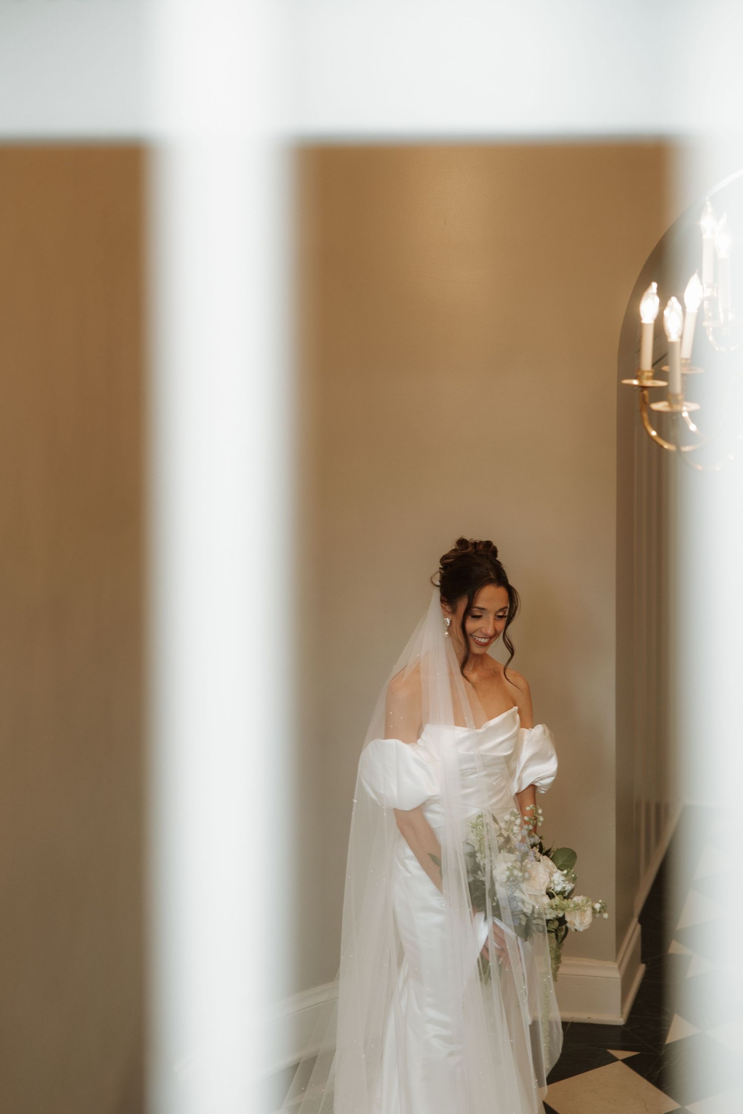 A bride in a white wedding dress and veil walks down a hallway holding a bouquet of white flowers.