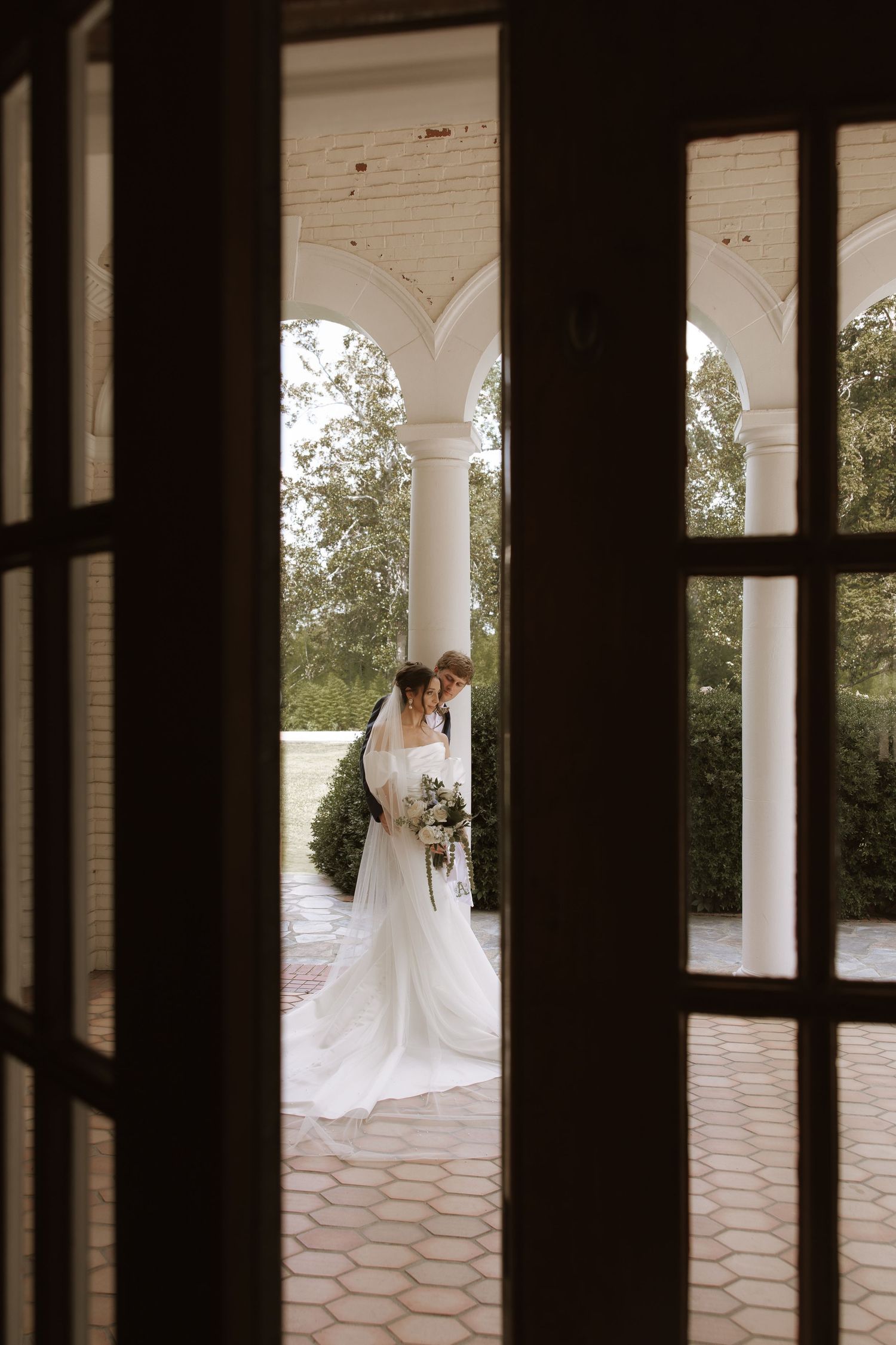 Elegant white wedding dress photographed through architectural columns on a covered porch with lush greenery in background.