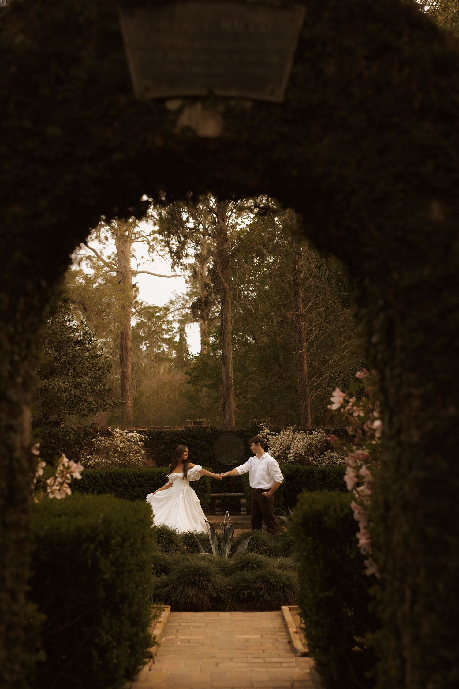 A bride and groom dance in a lush garden framed by a hedge archway at golden hour.