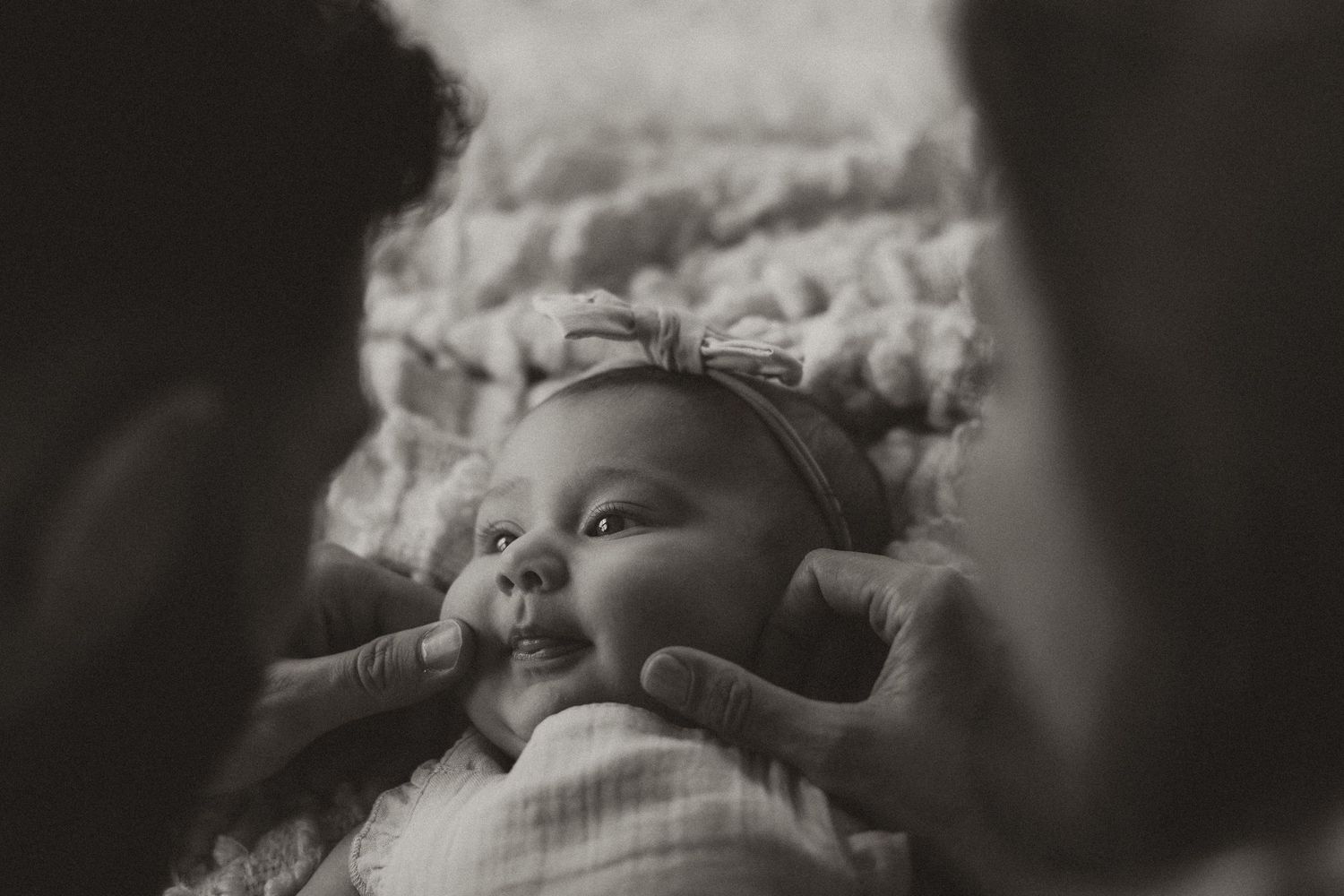 Black and white photo of a smiling newborn baby wearing a bow headband, gently touched by parents hands.