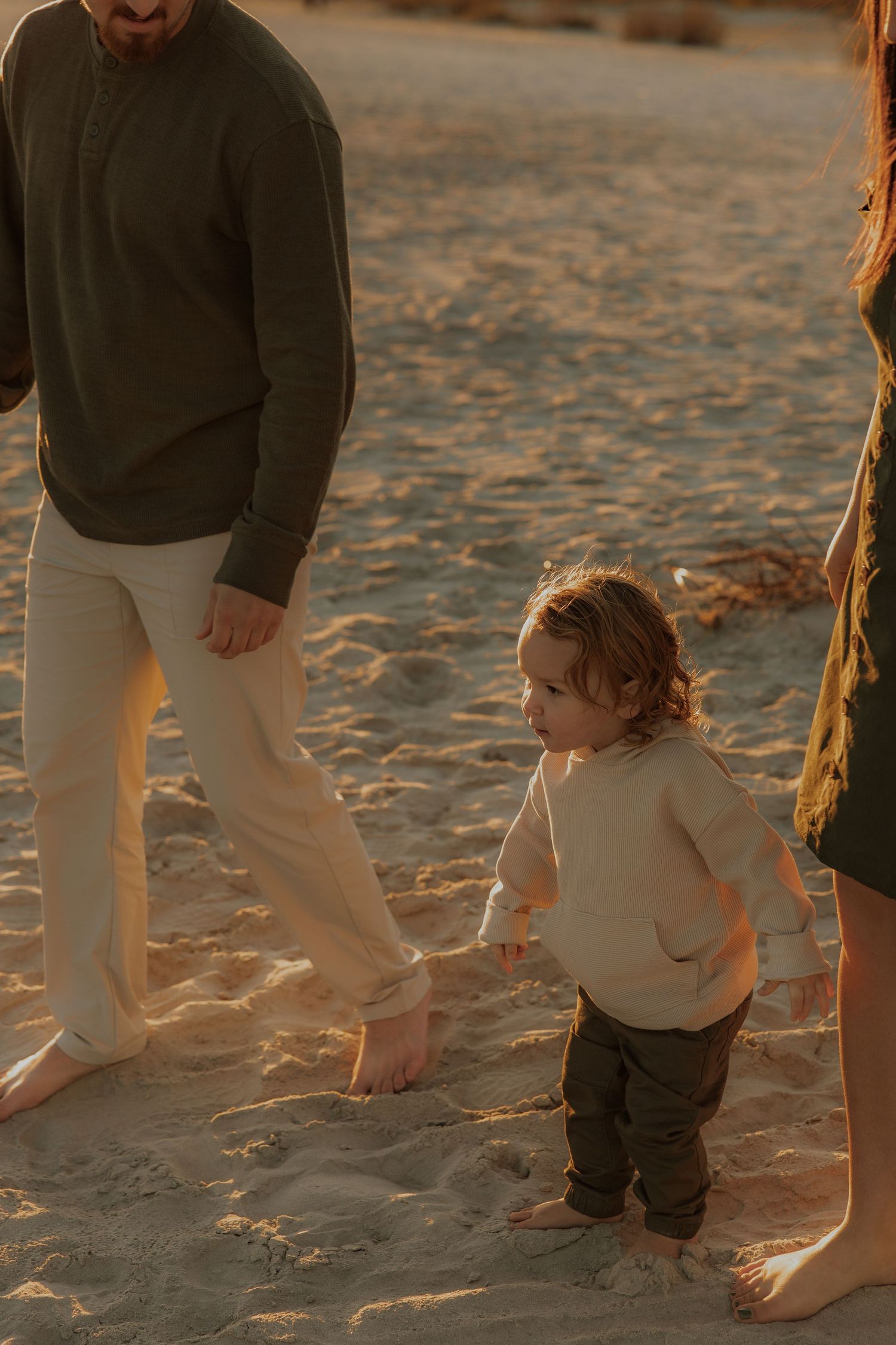 A toddler explores the beach sand at golden hour with adults nearby.