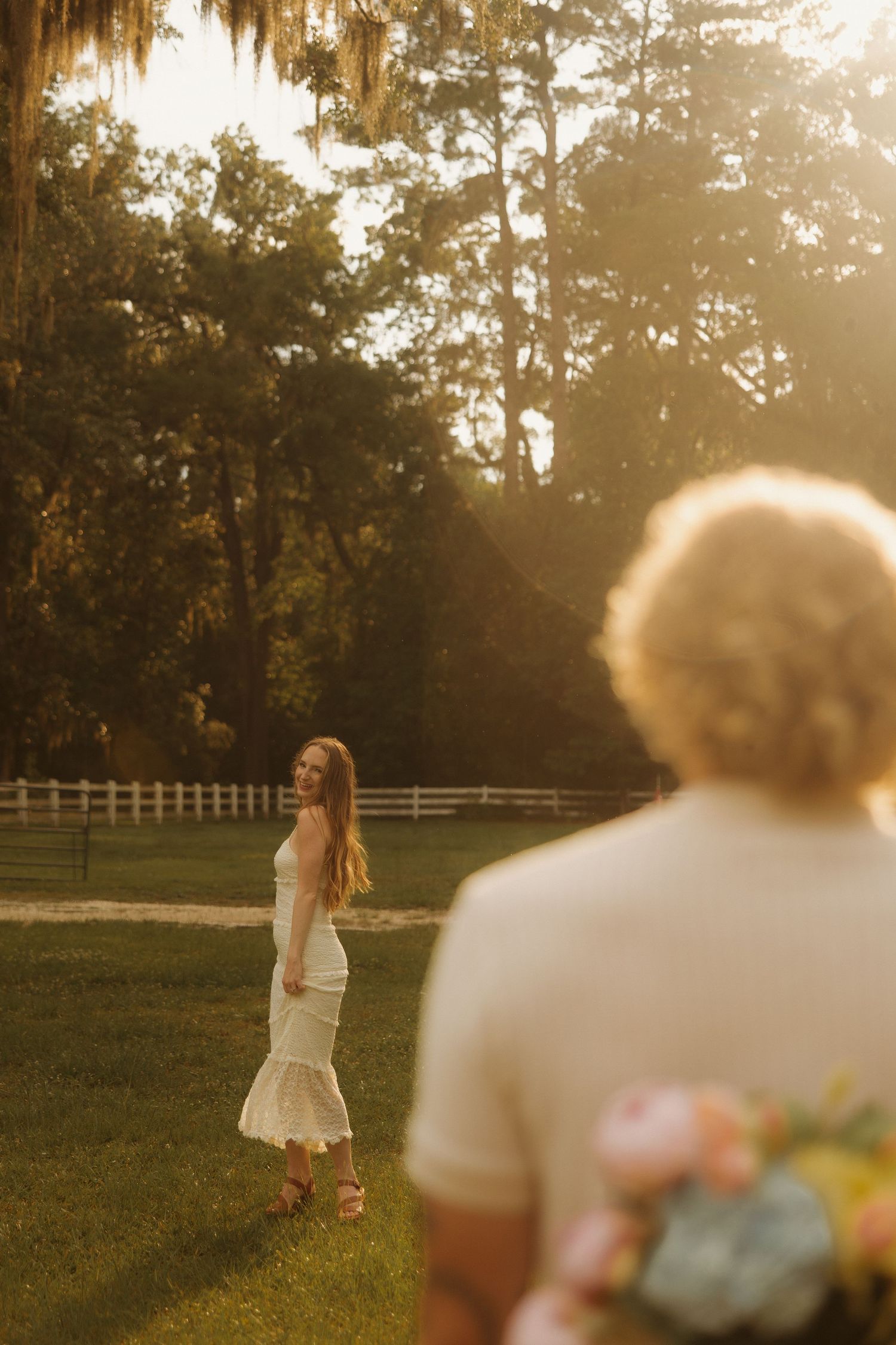 Golden hour photo of a bride in white dress standing on green grass with groom holding flowers in soft bokeh foreground.