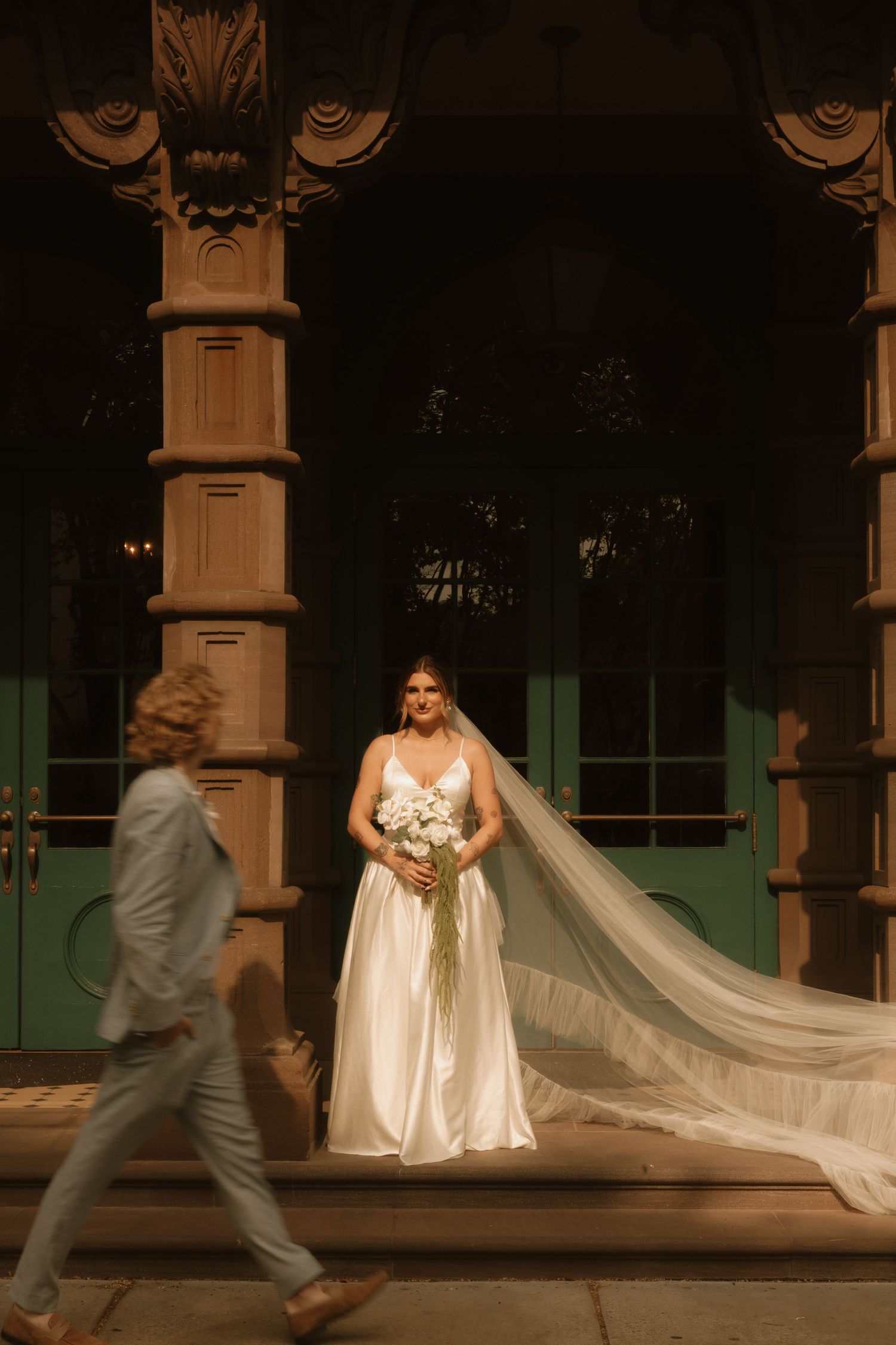 Bride in white gown and cathedral veil holds bouquet before ornate building entrance as suited figure walks past.