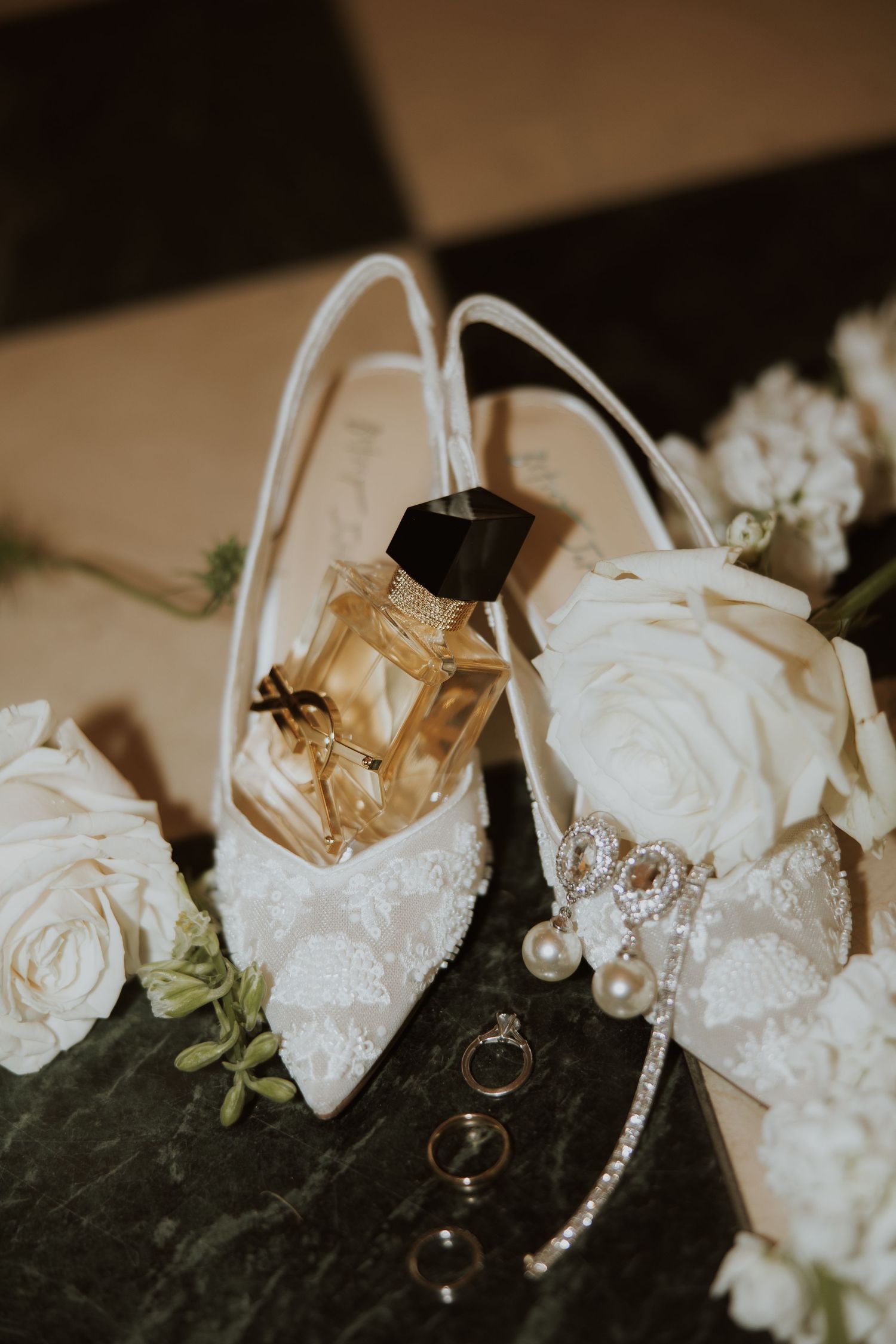 Wedding rings and white roses lay artistically arranged on a dark surface with soft sepia lighting.