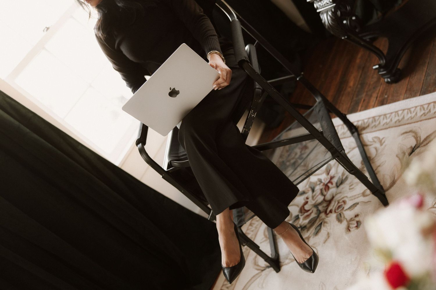 Professional woman in black outfit holding a MacBook laptop while seated on a chair in an elegant office setting.