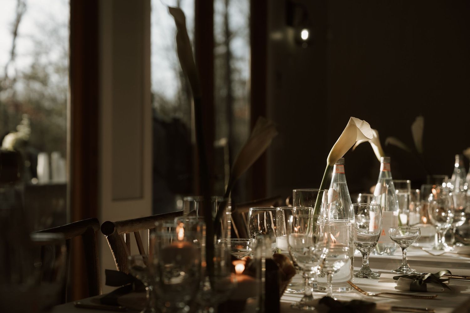 Elegant dinner table setting with crystal glasses, candles, and dried floral centerpieces in a moody, dimly lit dining room.