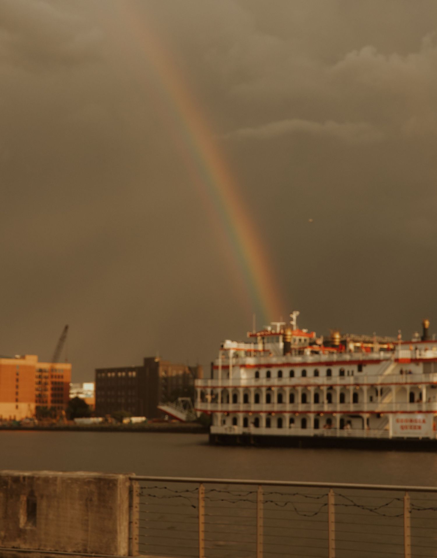 A dramatic double rainbow arches over a waterfront with boats and buildings silhouetted against a moody evening sky.