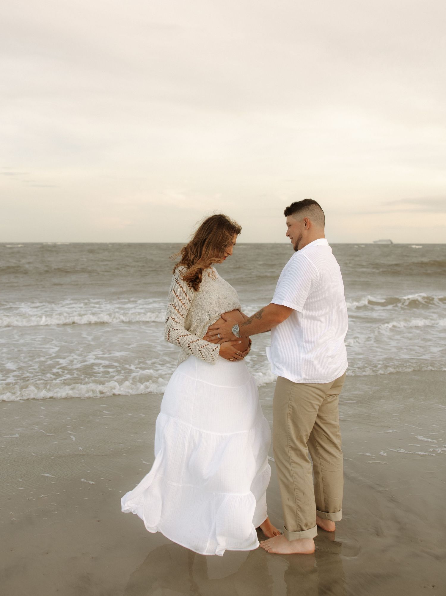 Romantic couple dancing and twirling together on a misty beach shoreline at sunset in coordinated white and beige outfits.