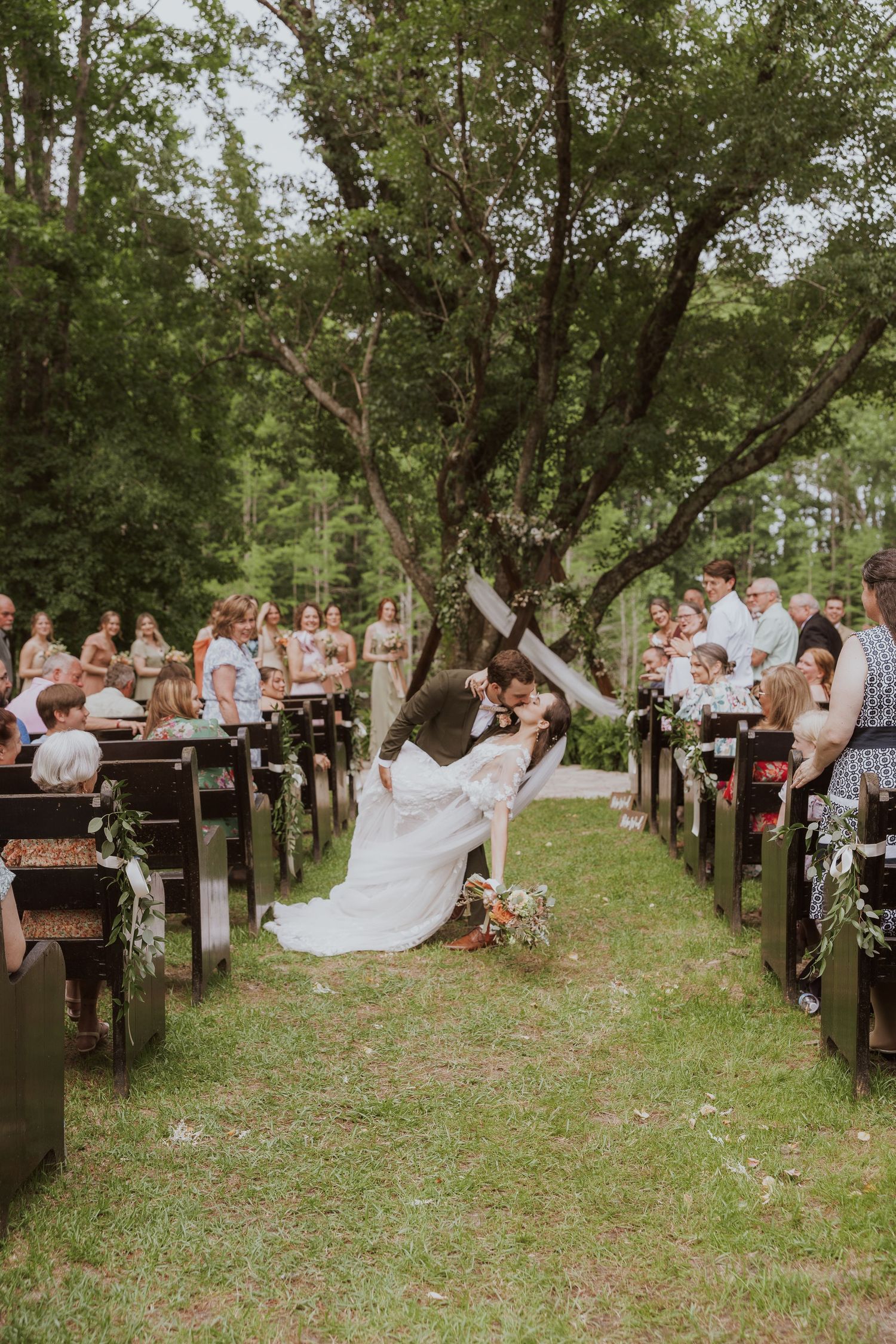 A series of outdoor wooden benches arranged in rows for a garden wedding ceremony with guests seated under green trees.