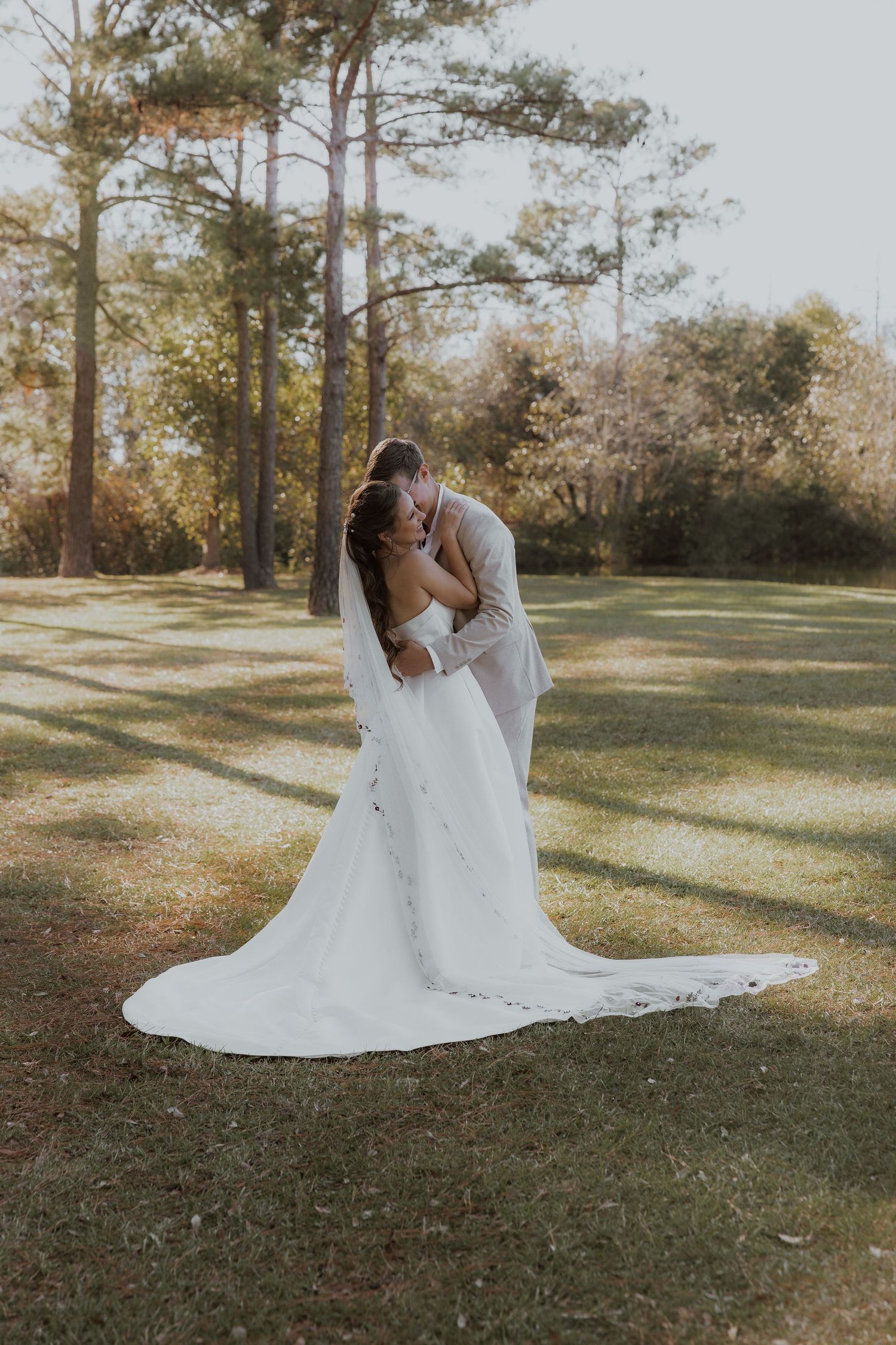 A romantic wedding photo on a sunlit lawn with pine trees in the background.