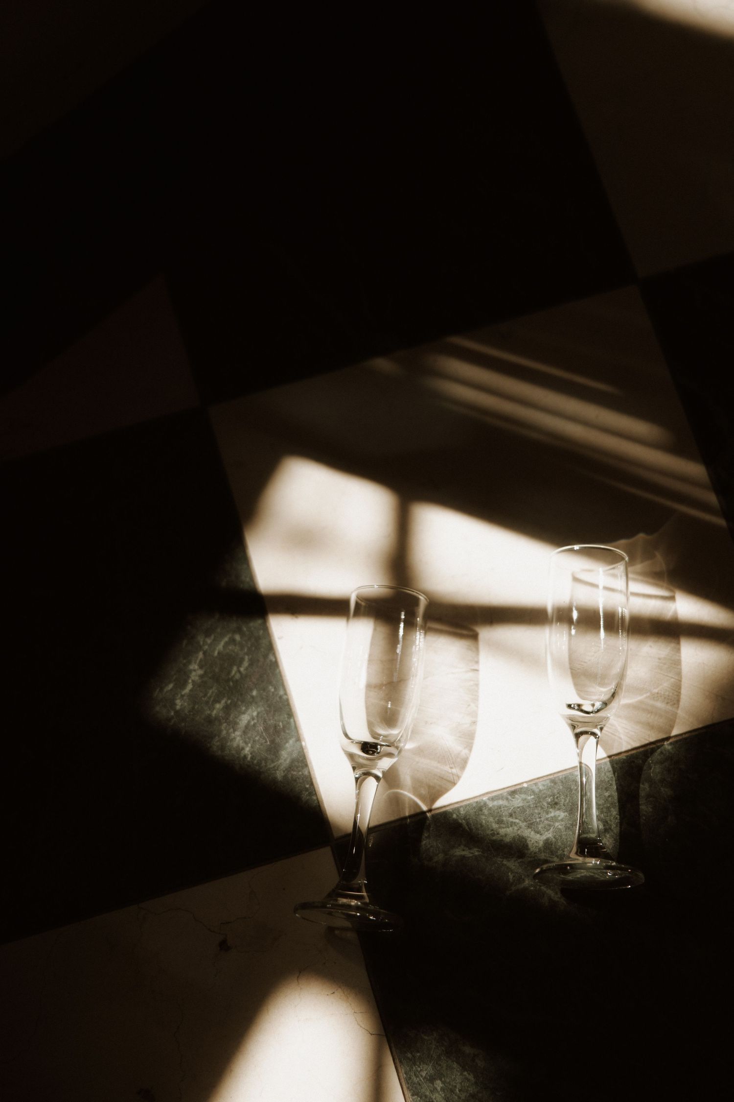 Dramatic sepia-toned photograph of wine glasses casting long shadows in angular light and dark patterns.