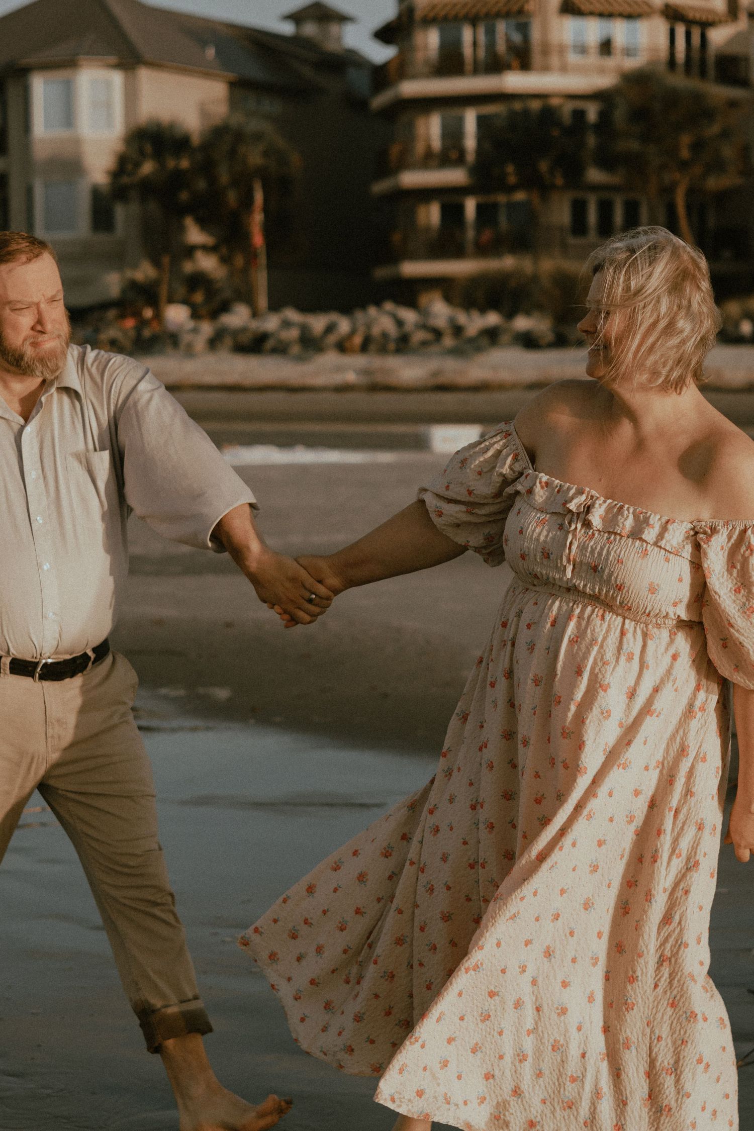 Couple dancing on the beach in casual attire with beach houses visible in the background.