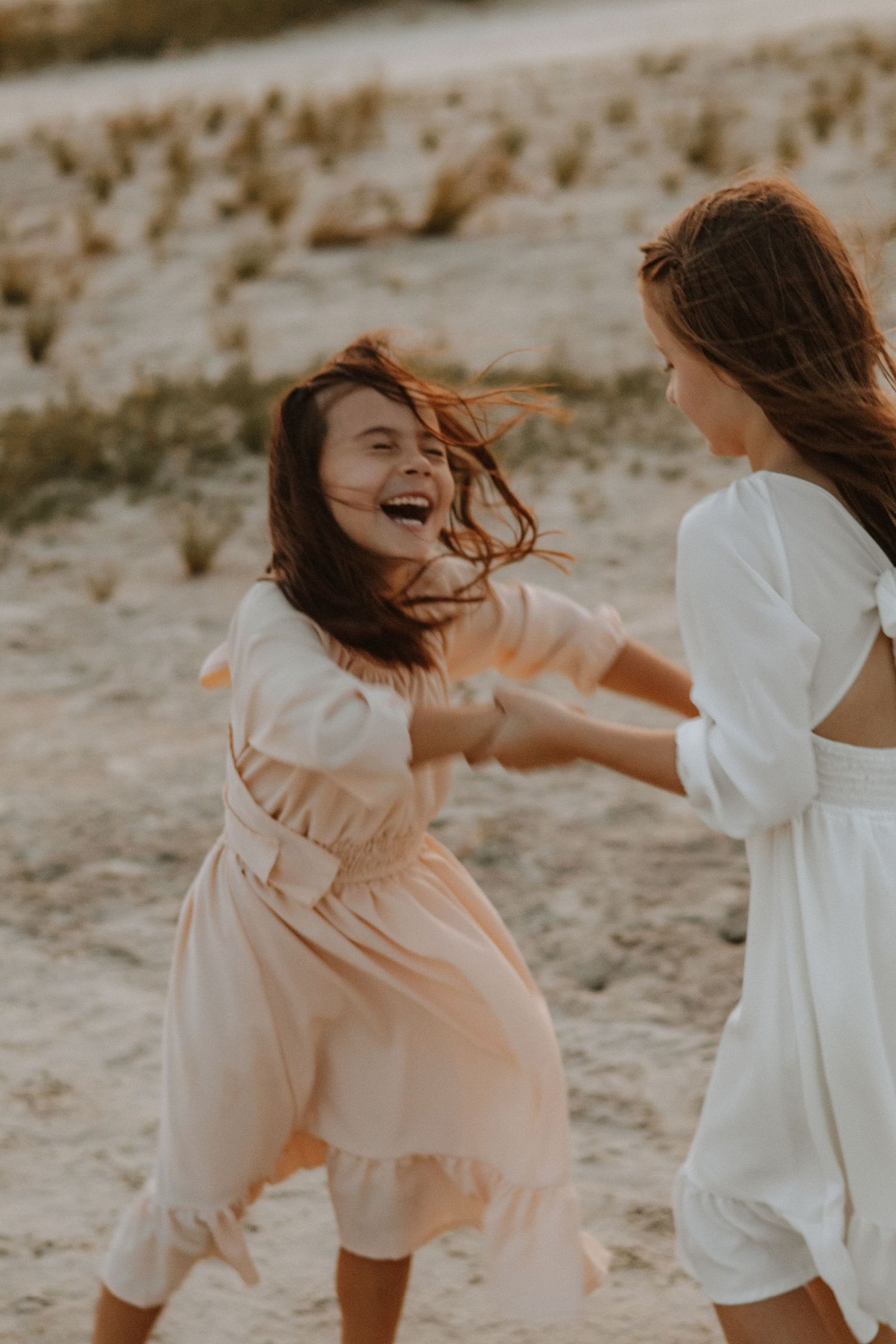 Two friends in flowy dresses dance and laugh together on a beach during golden hour.