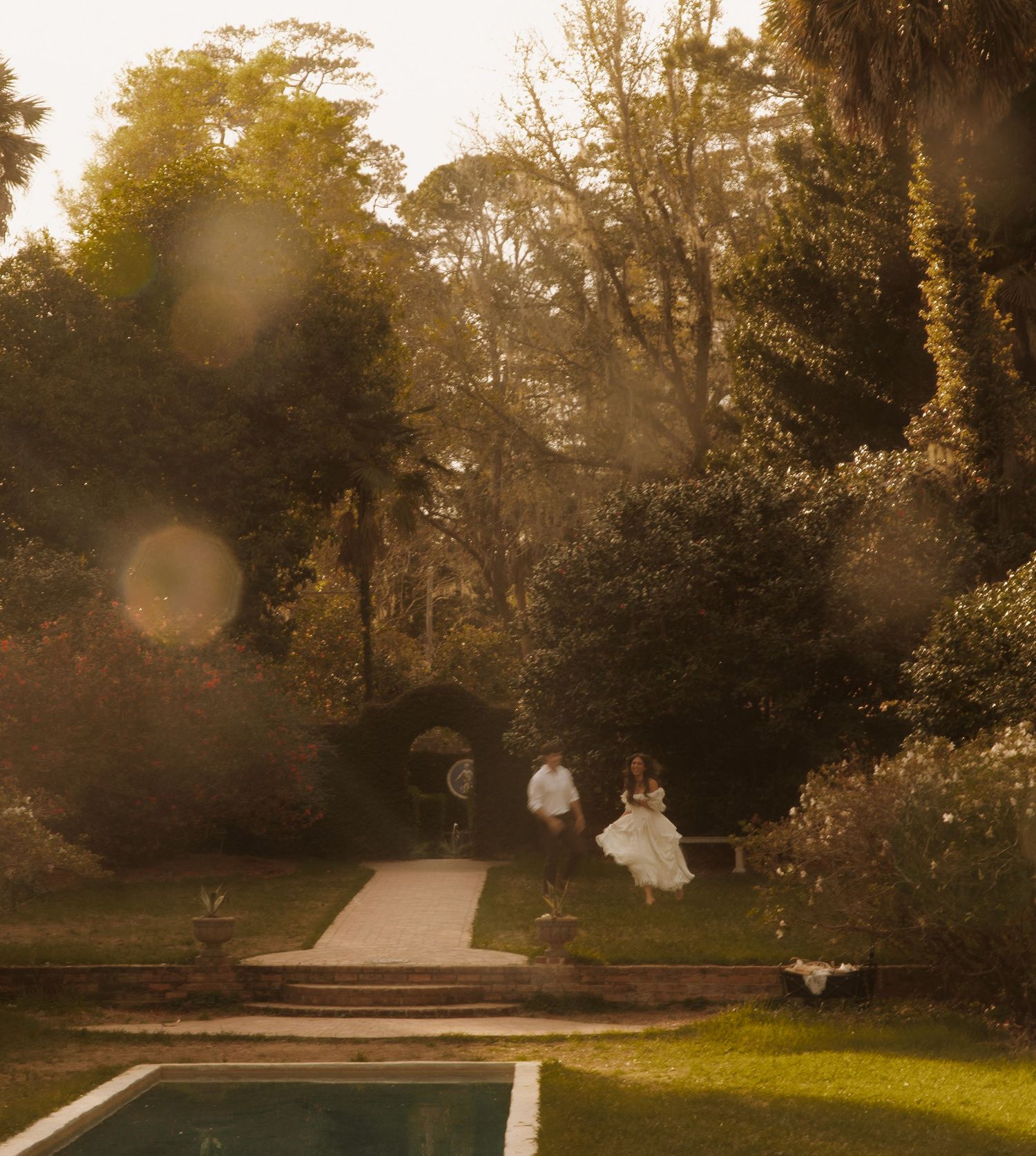 Couple in wedding attire running through a golden sunlit garden with lush trees and a pool in the foreground.