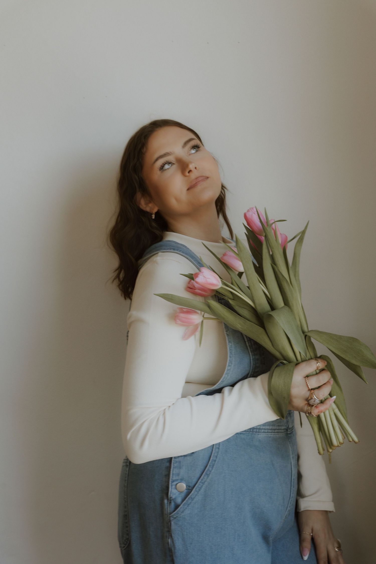 Person in denim overalls and white sweater holding pink tulips against a light wall.