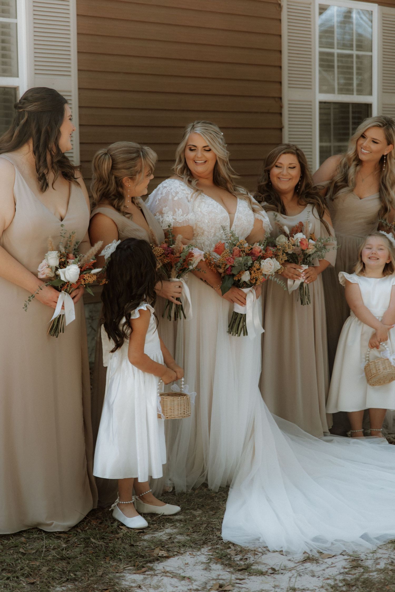 Bridal party in neutral dresses gathered around bride in white gown holding floral bouquets.