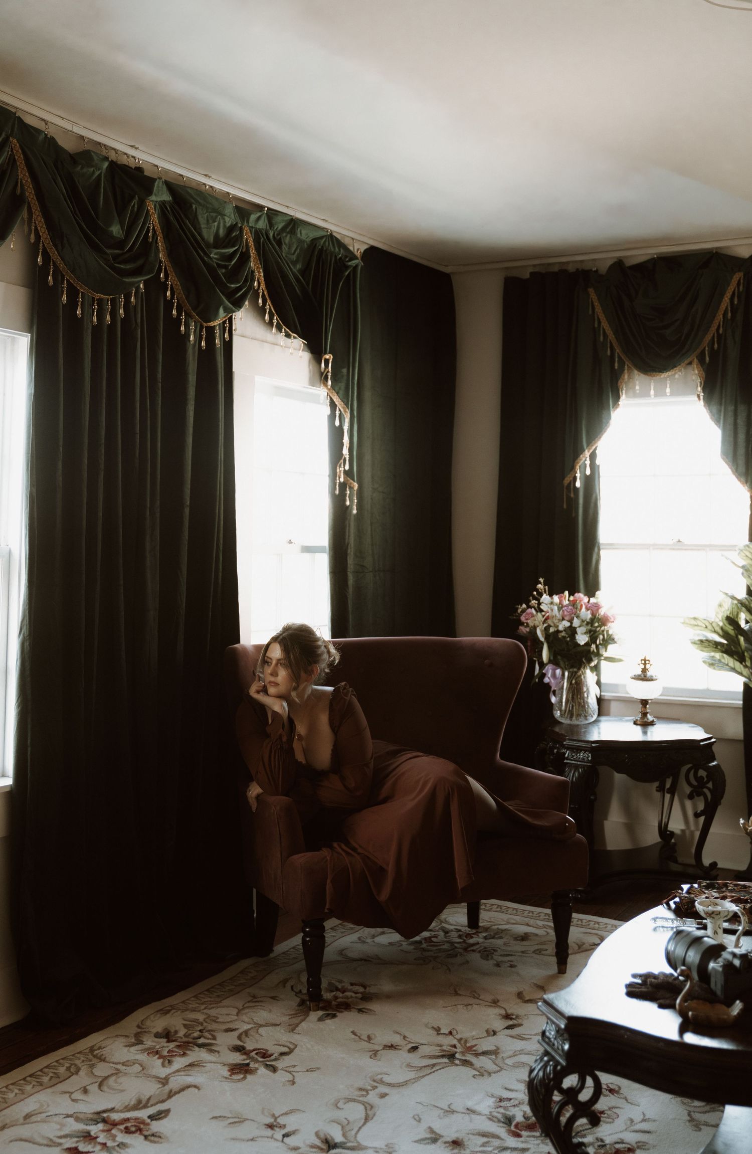 Elegant living room interior with dark curtains, upholstered armchair, and floral carpet in natural lighting.