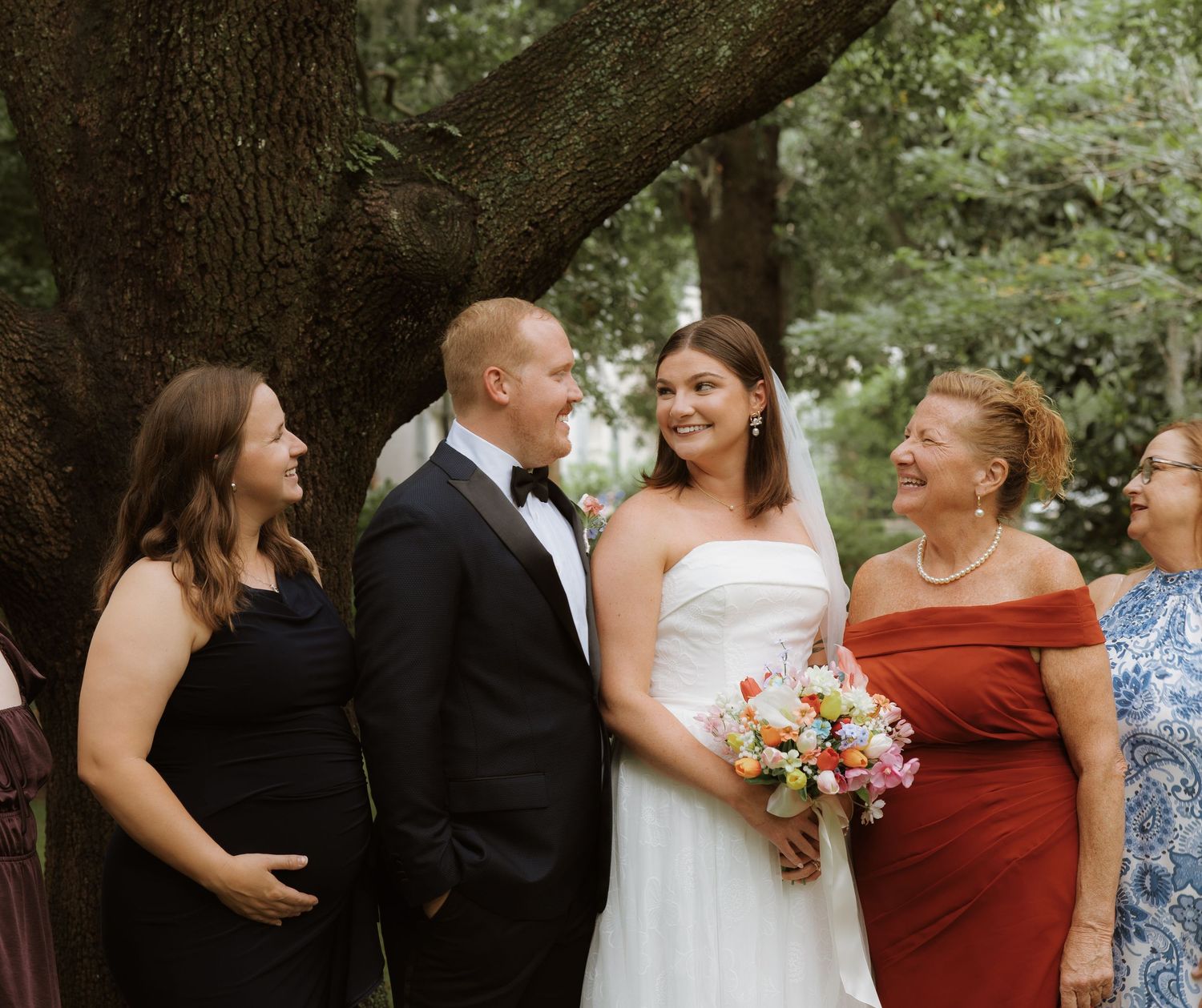 Group of wedding party members in formal attire gathered outdoors with bridesmaids in burgundy dresses.