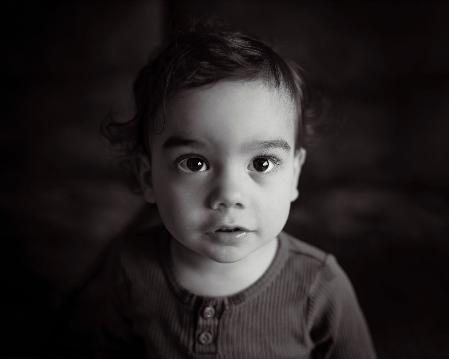 A black and white portrait of a young child with dark hair wearing a henley shirt against a dark background.