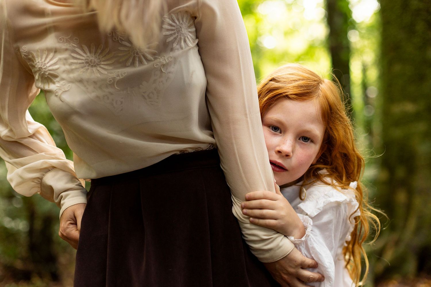 A young child with red hair clings to someone's arm while standing in a sunlit forest setting.