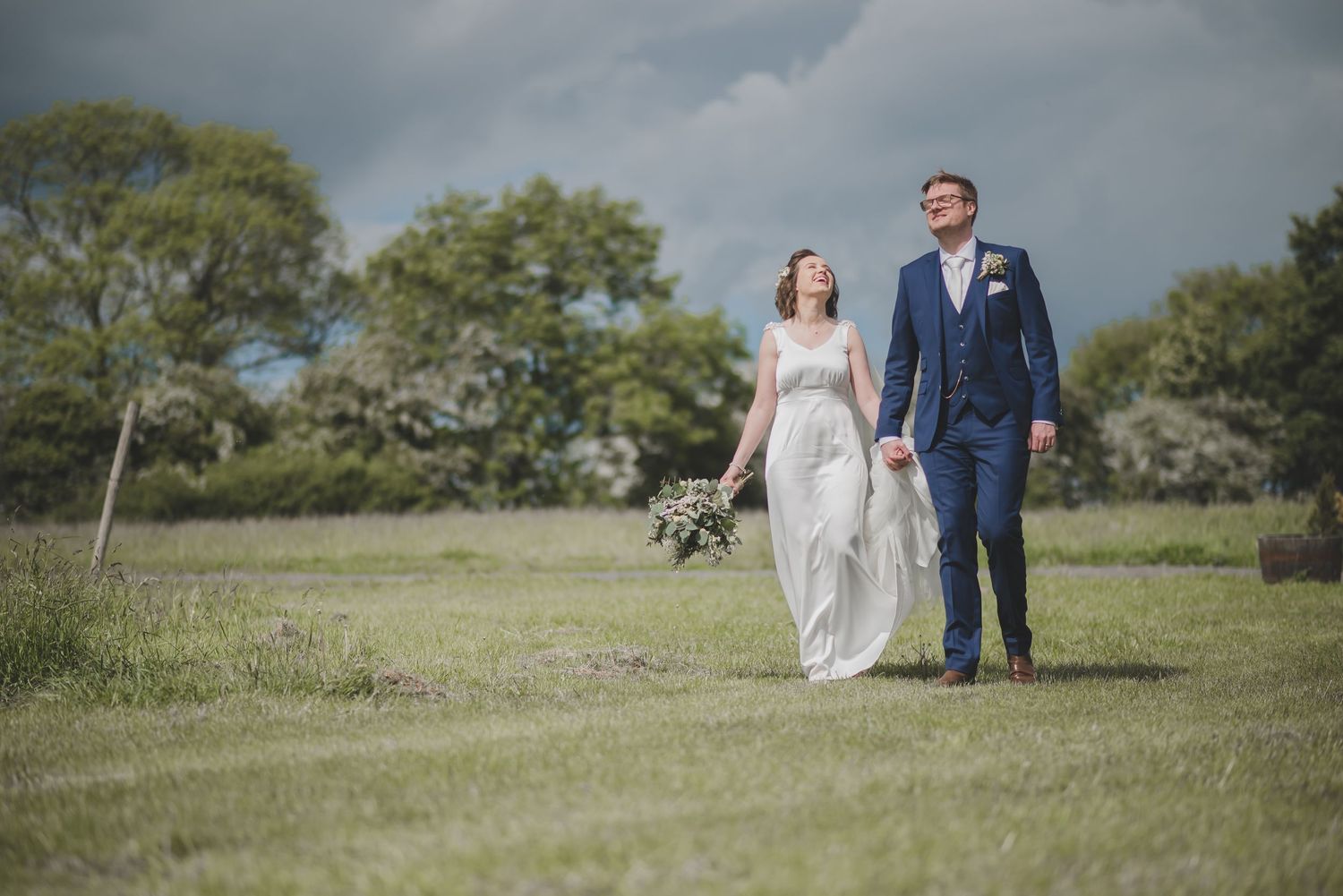 A bride and groom walk together across a green field on their wedding day with trees in the background.