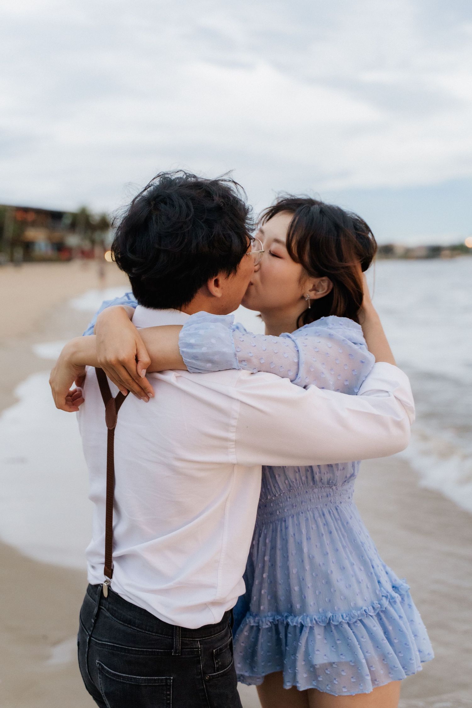 Romantic couple embracing and kissing on a beach during sunset with water and buildings in the background.