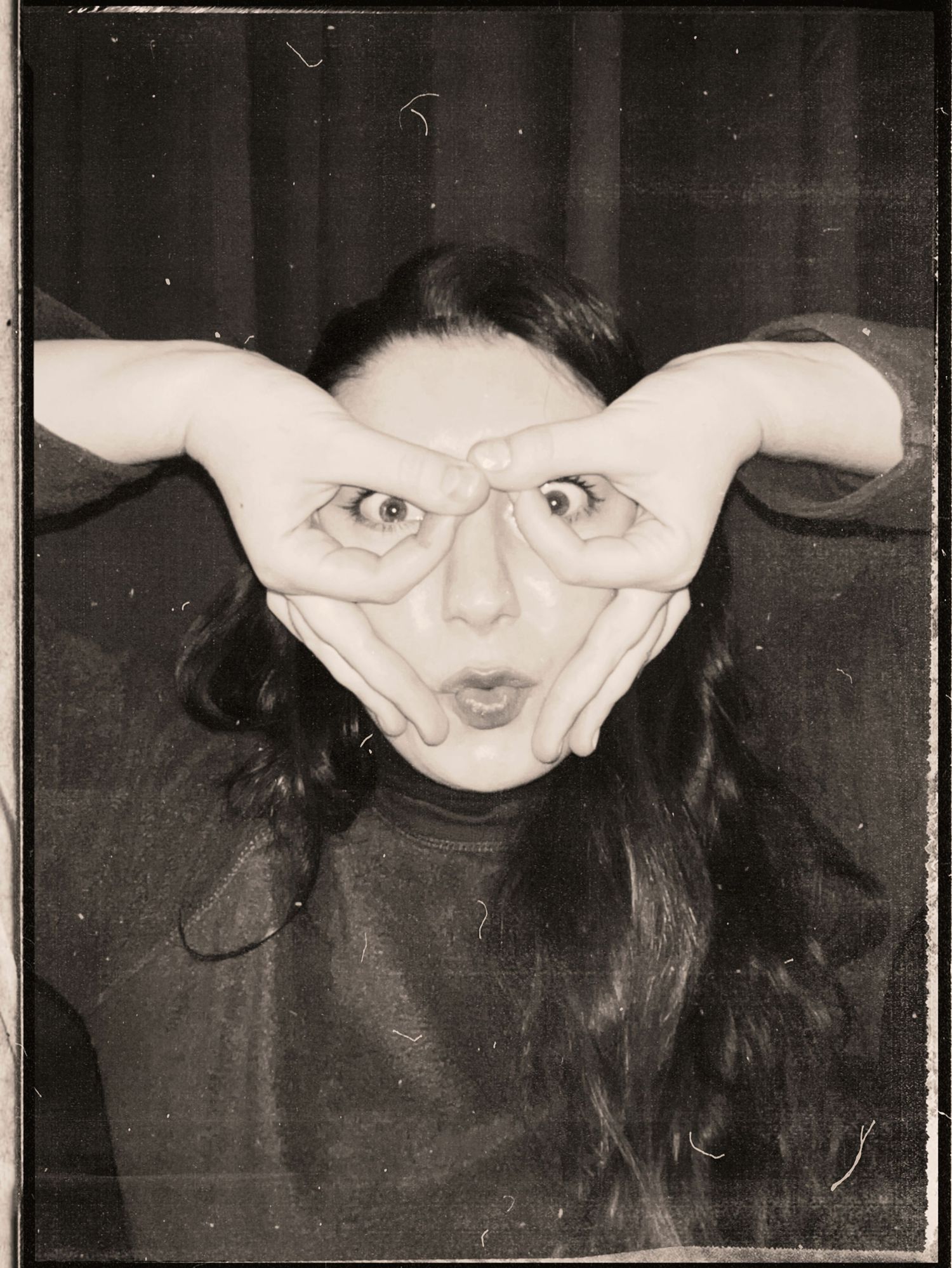 Black and white photo booth image of a woman making a funny face with hands framing her wide eyes.