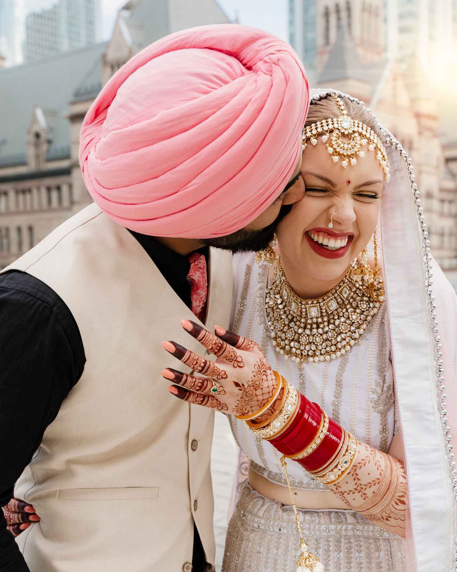 A joyful moment between a bride in traditional Indian wedding attire and groom in pink turban sharing an intimate moment.