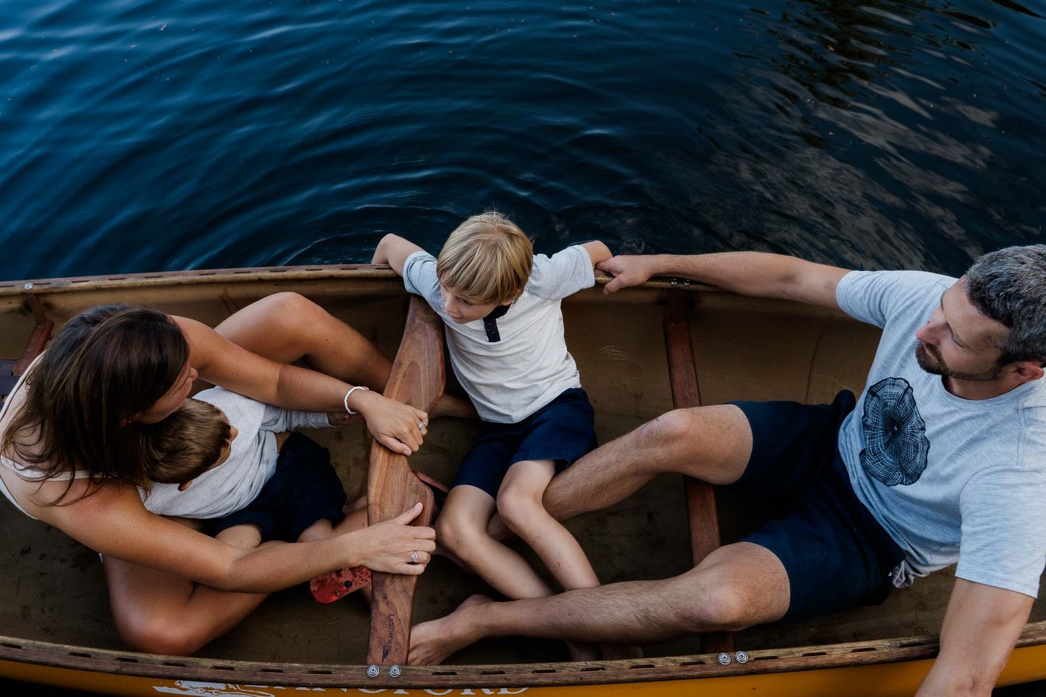 Family relaxing together in wooden canoe on calm blue water during summer day.