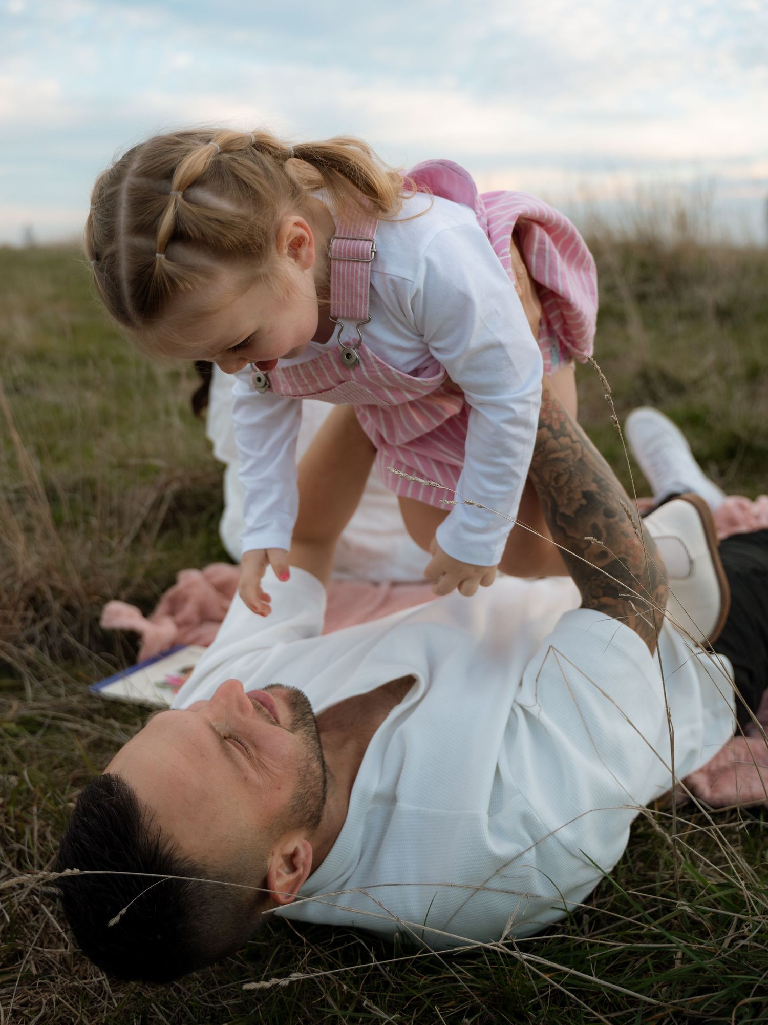A father lies in a grassy field lifting his laughing toddler daughter wearing pink overalls above him.