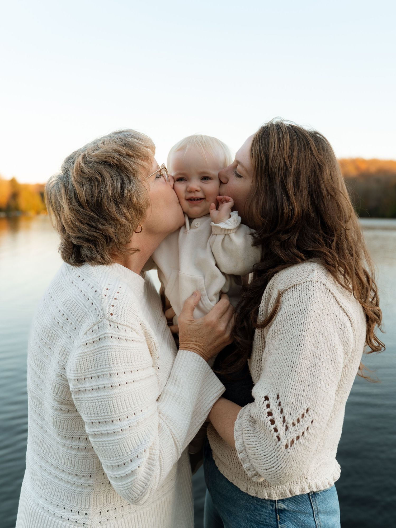Two women in white sweaters kiss a smiling baby by a lake at golden hour.