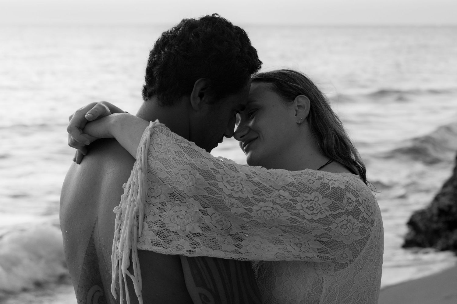 Two figures share an intimate moment by the ocean shore in black and white photography.