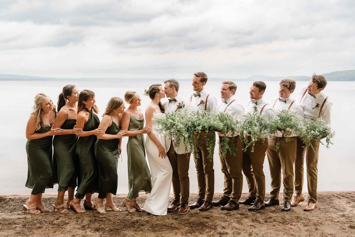 Wedding party stands together on beach with bridesmaids in dark dresses and groomsmen holding bouquets.