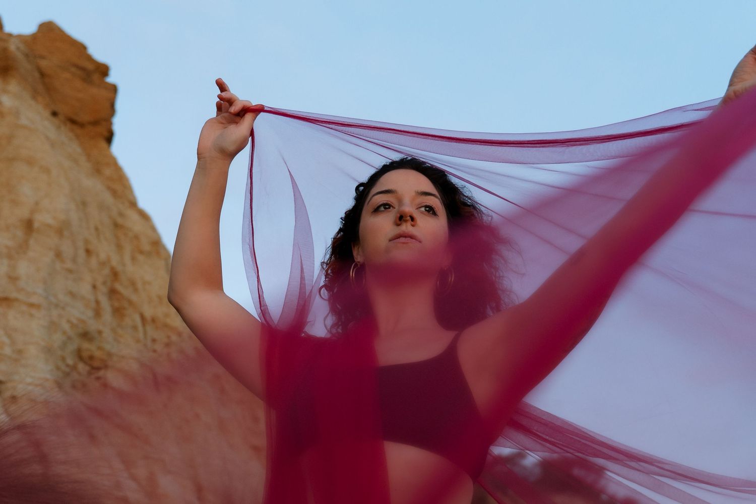 A figure holds up a flowing pink fabric against a desert landscape with orange rock formations at sunset.