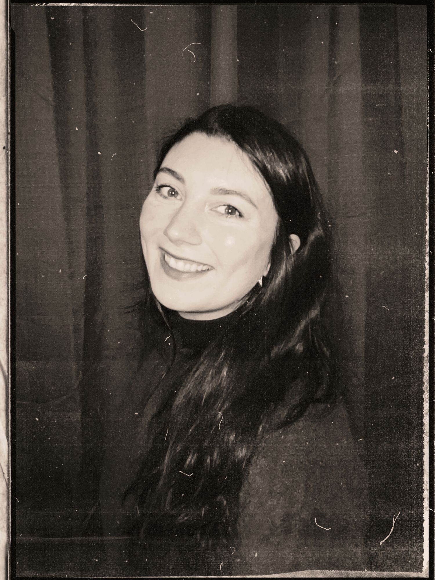 Vintage black and white photo booth portrait of a young woman with long dark hair, smiling warmly.