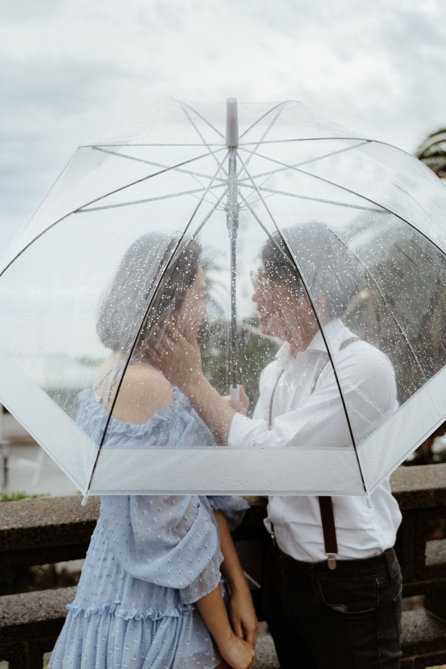 A couple shares an intimate moment under a clear umbrella on an overcast day.