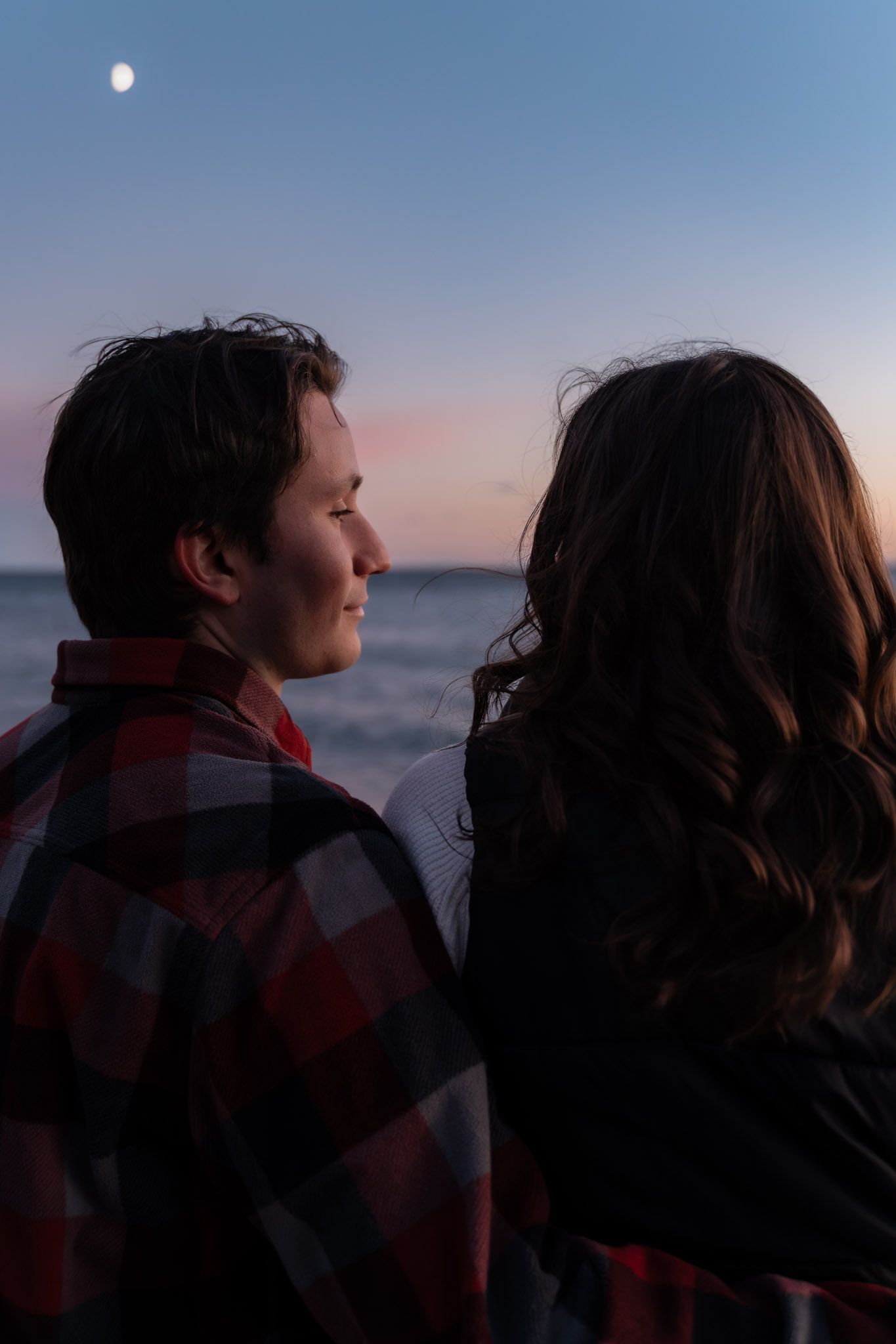 Silhouetted couple in plaid shirts watching sunset over ocean with moon visible.