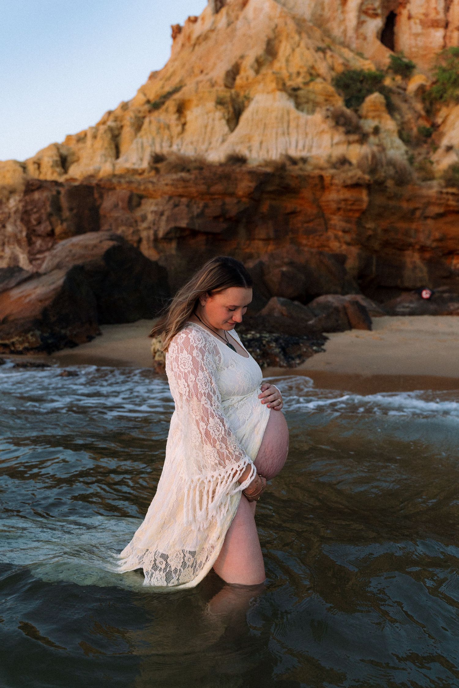 White lace maternity dress photoshoot at sunset on a beach with dramatic cliffs and caves in the background.