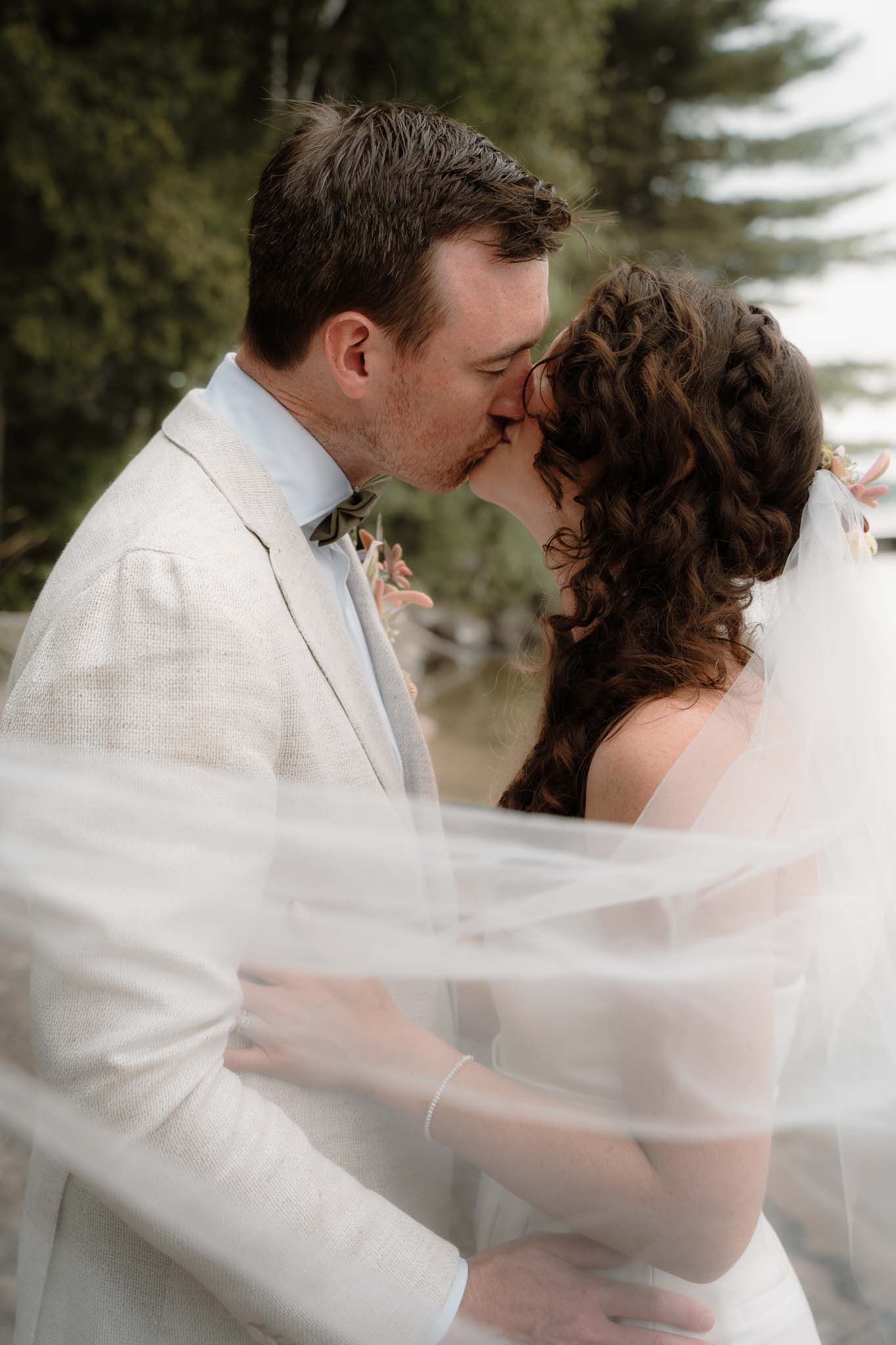 Intimate wedding moment captured with flowing veil and white suit outdoors.