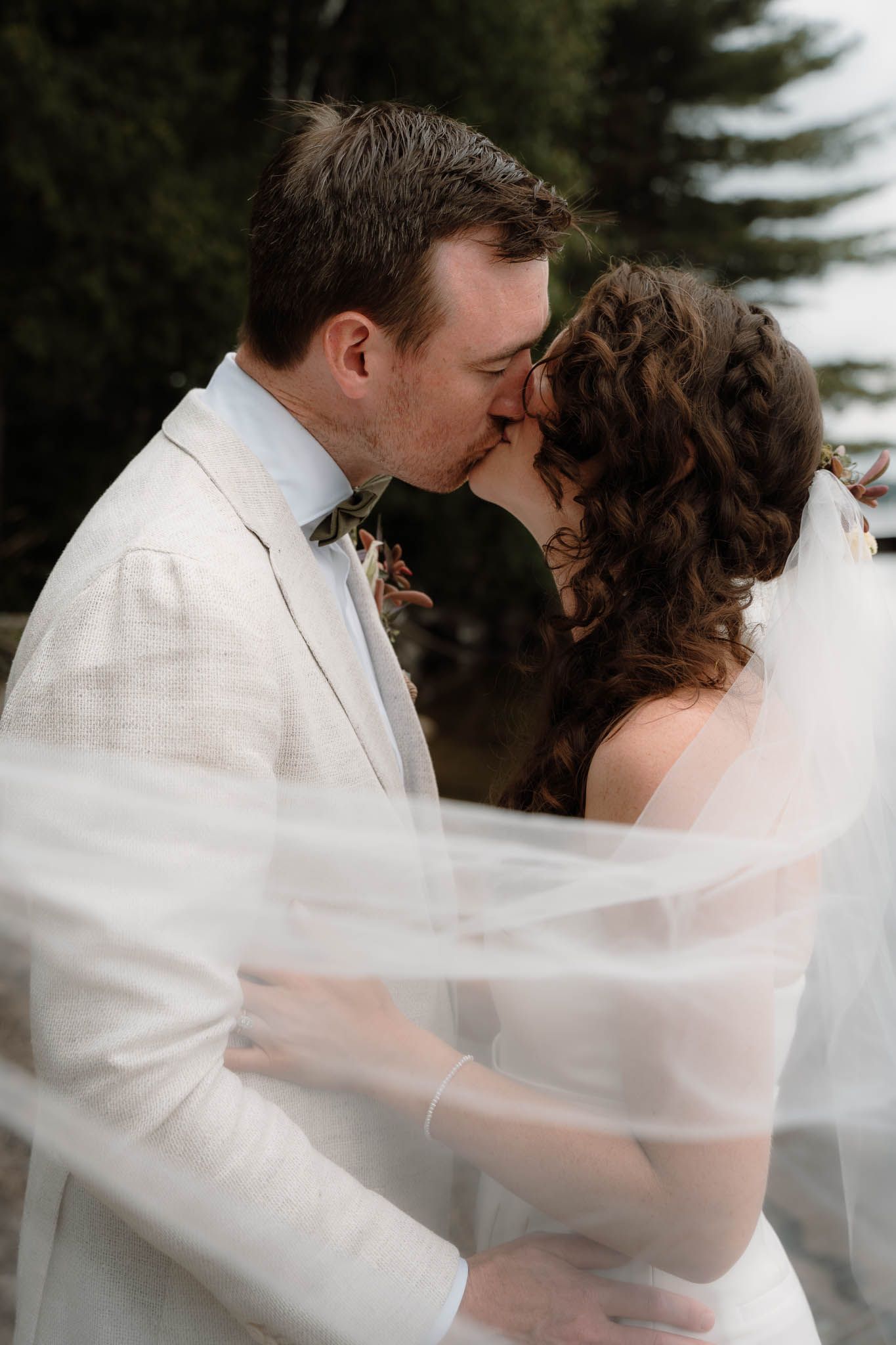 Romantic newlywed couple share a kiss outdoors with flowing veil and white suit.
