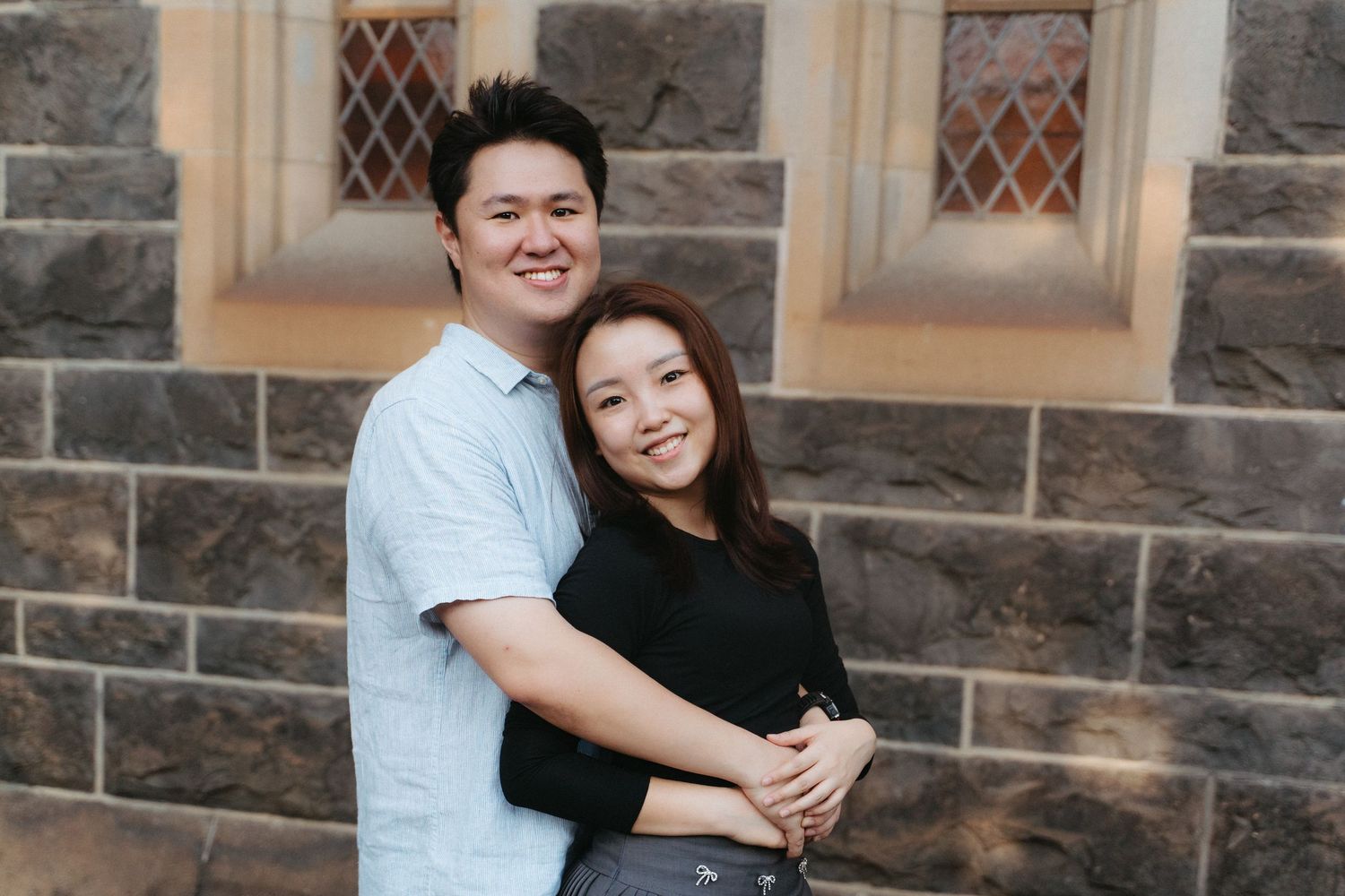 A couple embraces while smiling together in front of a stone building with diamond-paned windows.