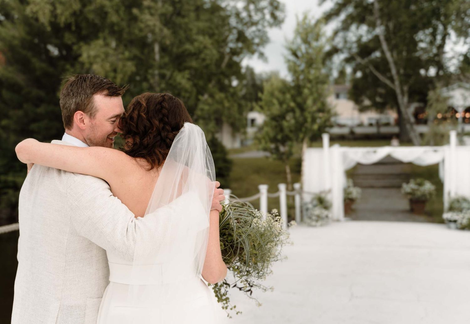 Couple in white embracing at outdoor wedding venue with garden path.