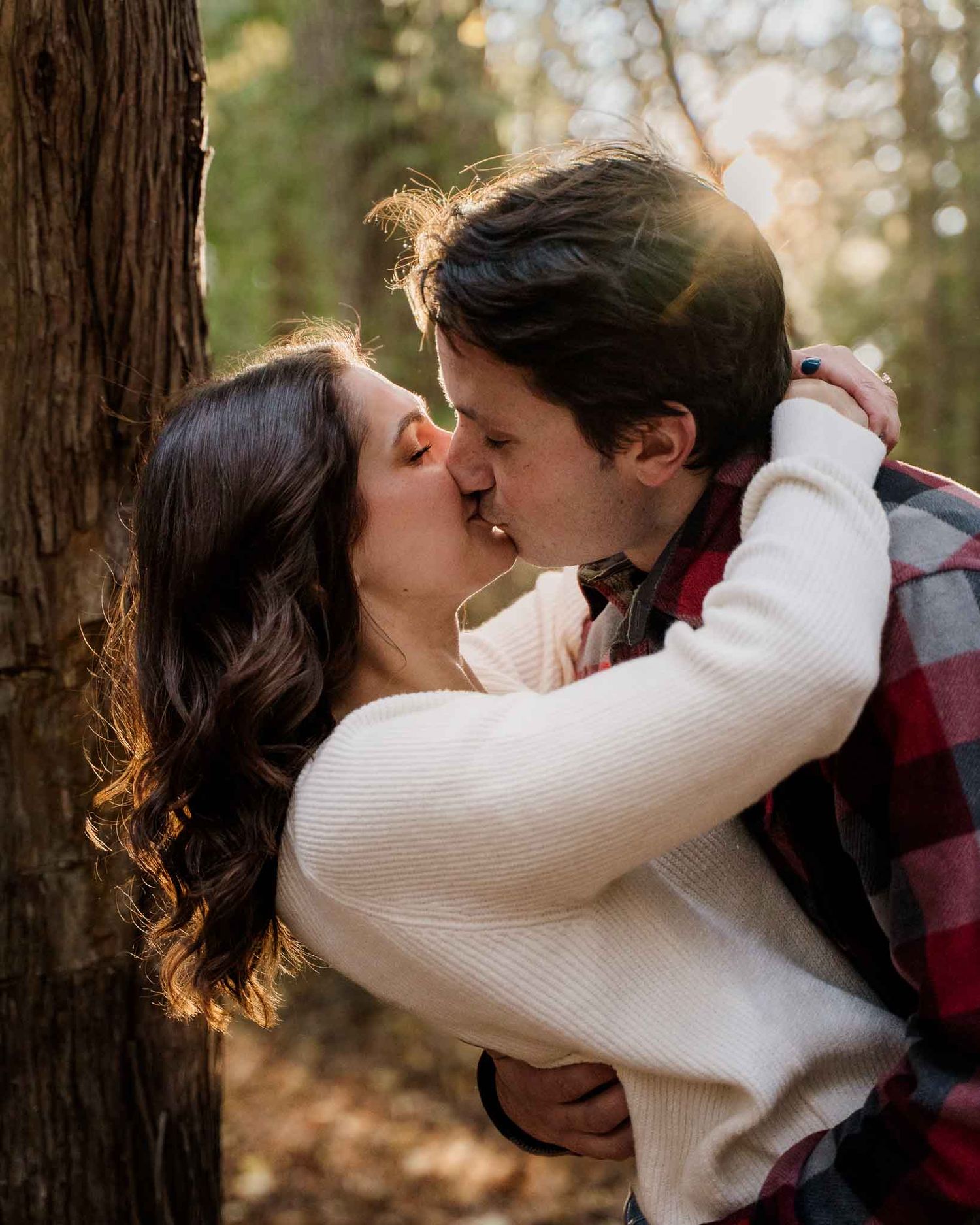 A romantic couple sharing a kiss in front of a tree during golden hour in a natural outdoor setting.