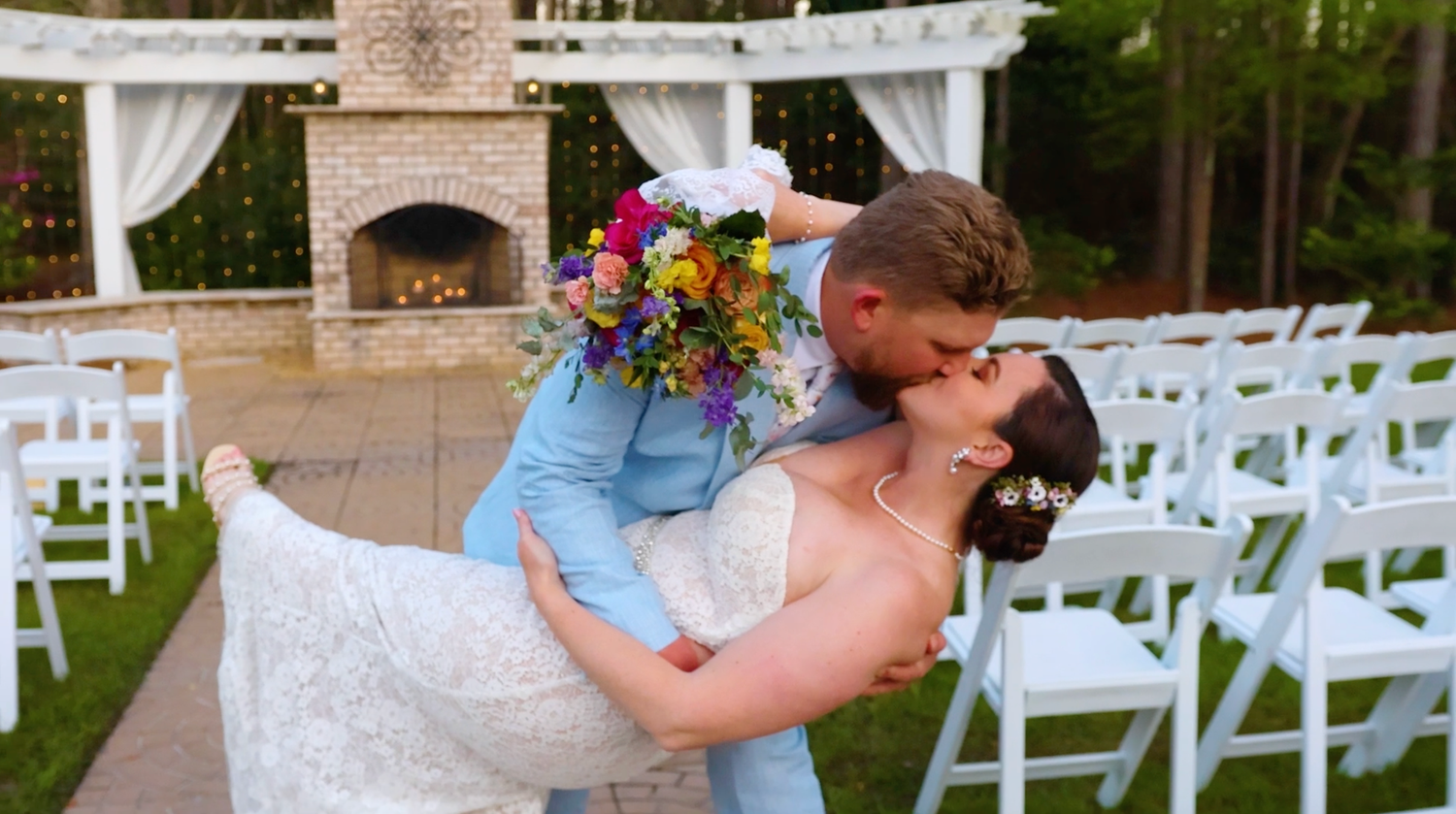 A romantic dance dip at an outdoor garden wedding ceremony setup with white chairs and floral arrangements.