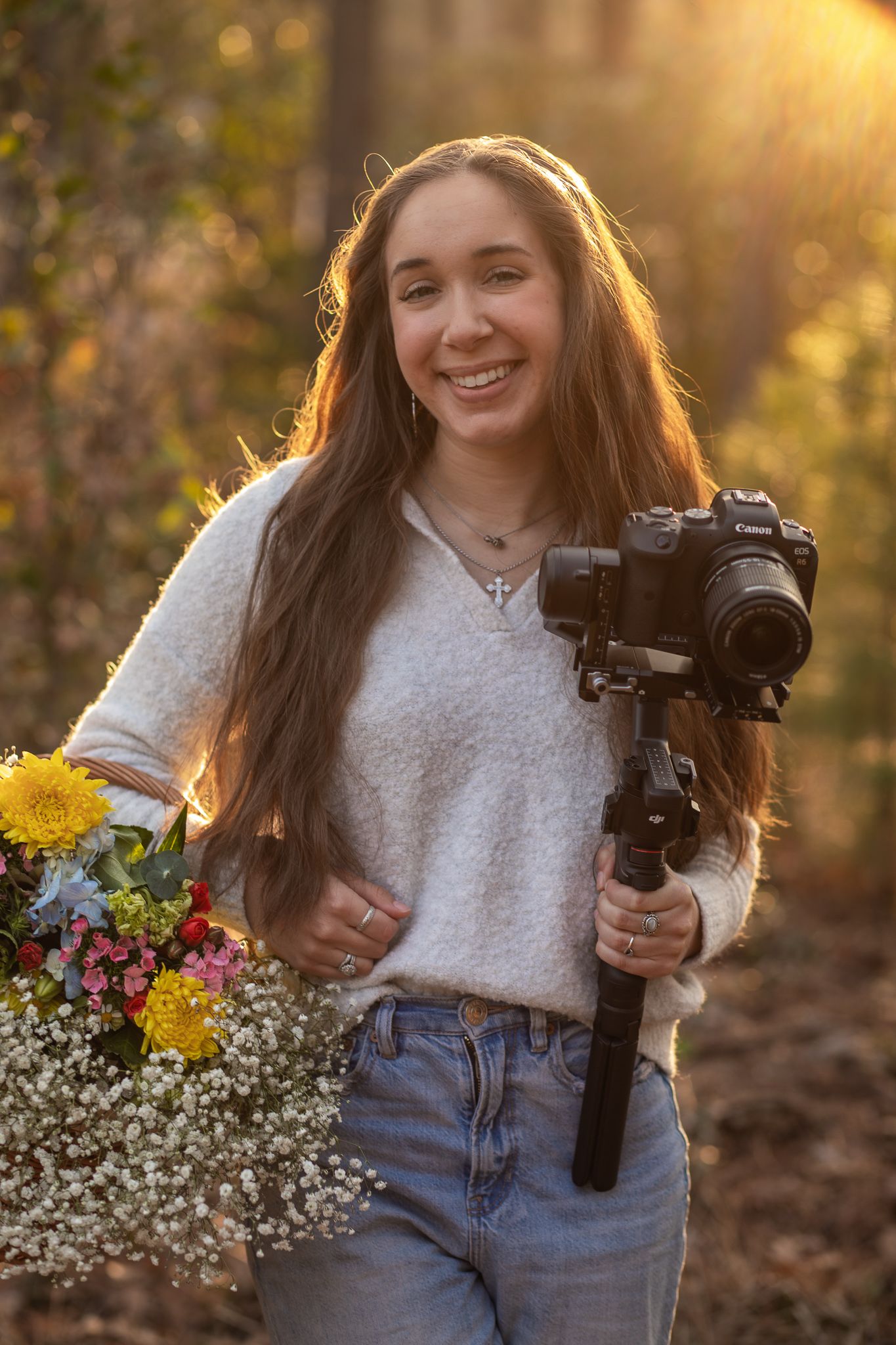 Photographer holding camera gimbal and colorful bouquet of flowers in golden sunlight during outdoor photo session.