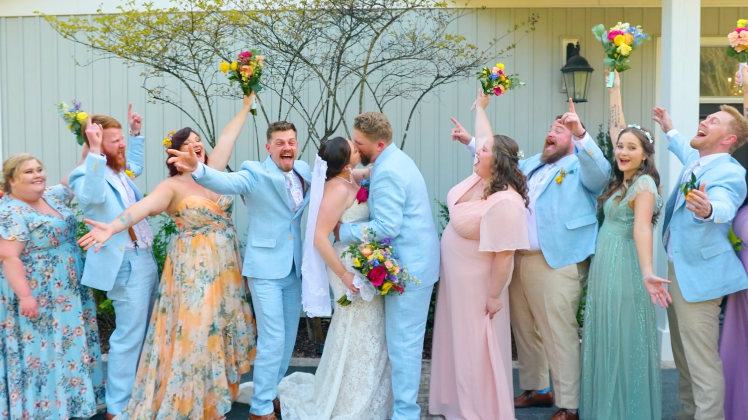 Wedding party in light blue suits and floral bridesmaid dresses pose for a fun group photo outdoors.